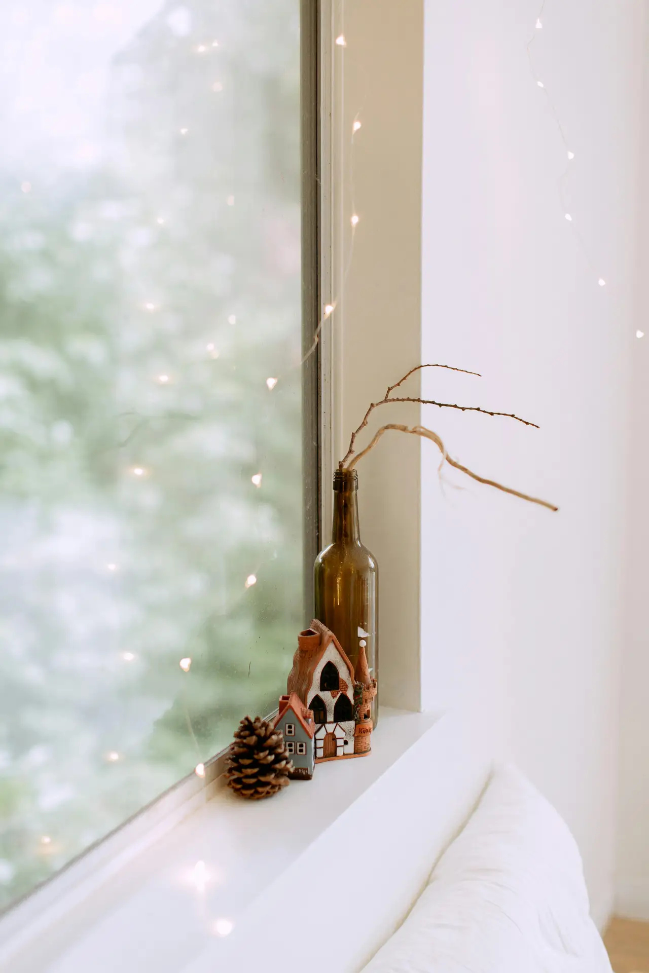 A serene, close-up shot of a white window ledge, illustrating simple small apartment decorating ideas. To decorate the windowsill, a green glass bottle repurposed as a vase holds a few slender, dried branches. Next to it sit two miniature ceramic houses and a single pinecone, creating a charming winter-themed display. Soft fairy lights are draped against the windowpane, blurring into a warm glow against the out-of-focus green trees outside.