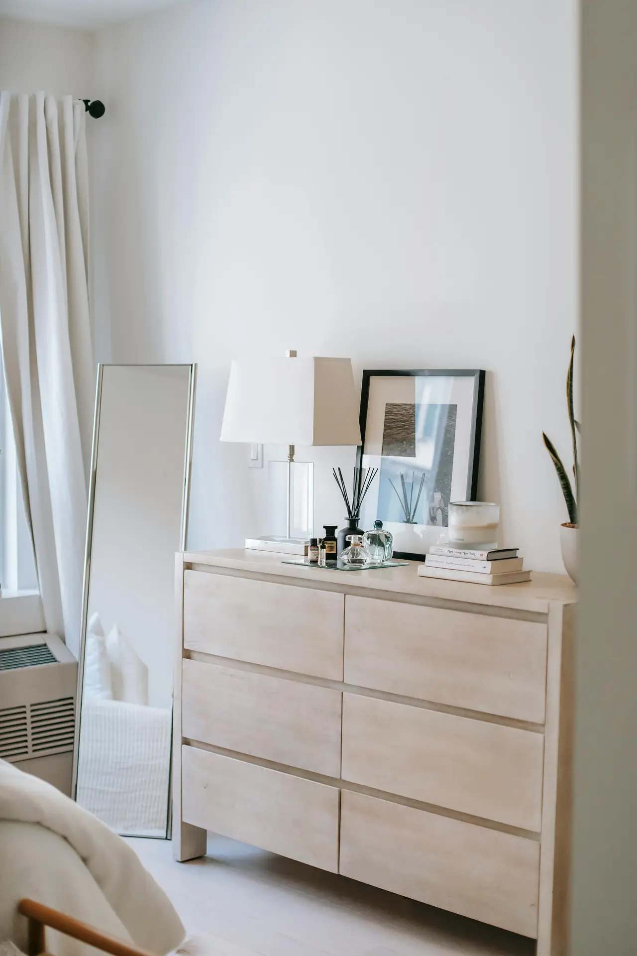 A bright and airy bedroom scene featuring elegant small apartment decorating ideas centered around a neutral-toned wooden dresser. The six-drawer dresser is styled with a white lamp, a black-framed art print, a reed diffuser, perfume bottles, and a small stack of books. To the left, a tall, frameless floor mirror leans against the white wall, reflecting a bit of the bed area. The minimalist aesthetic is enhanced by white curtains and a glimpse of a snake plant on the right, creating a serene and organized living space.