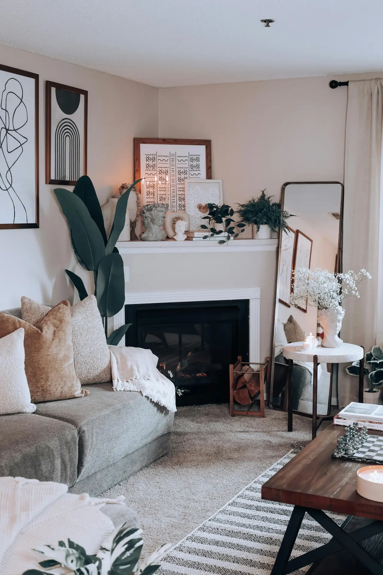 A cozy living room setting that provides inspiration for small apartment decorating ideas. The focal point is a corner fireplace with white mantel fireplace decor, featuring a collection of framed tribal and abstract art, bust sculptures, and small potted plants. A large leaning mirror reflects the room, while a grey sofa with plush velvet pillows and a wooden coffee table with a chess set complete the inviting atmosphere. A tall, dark leafy plant stands beside the fireplace, adding height and texture to the neutral-toned space.