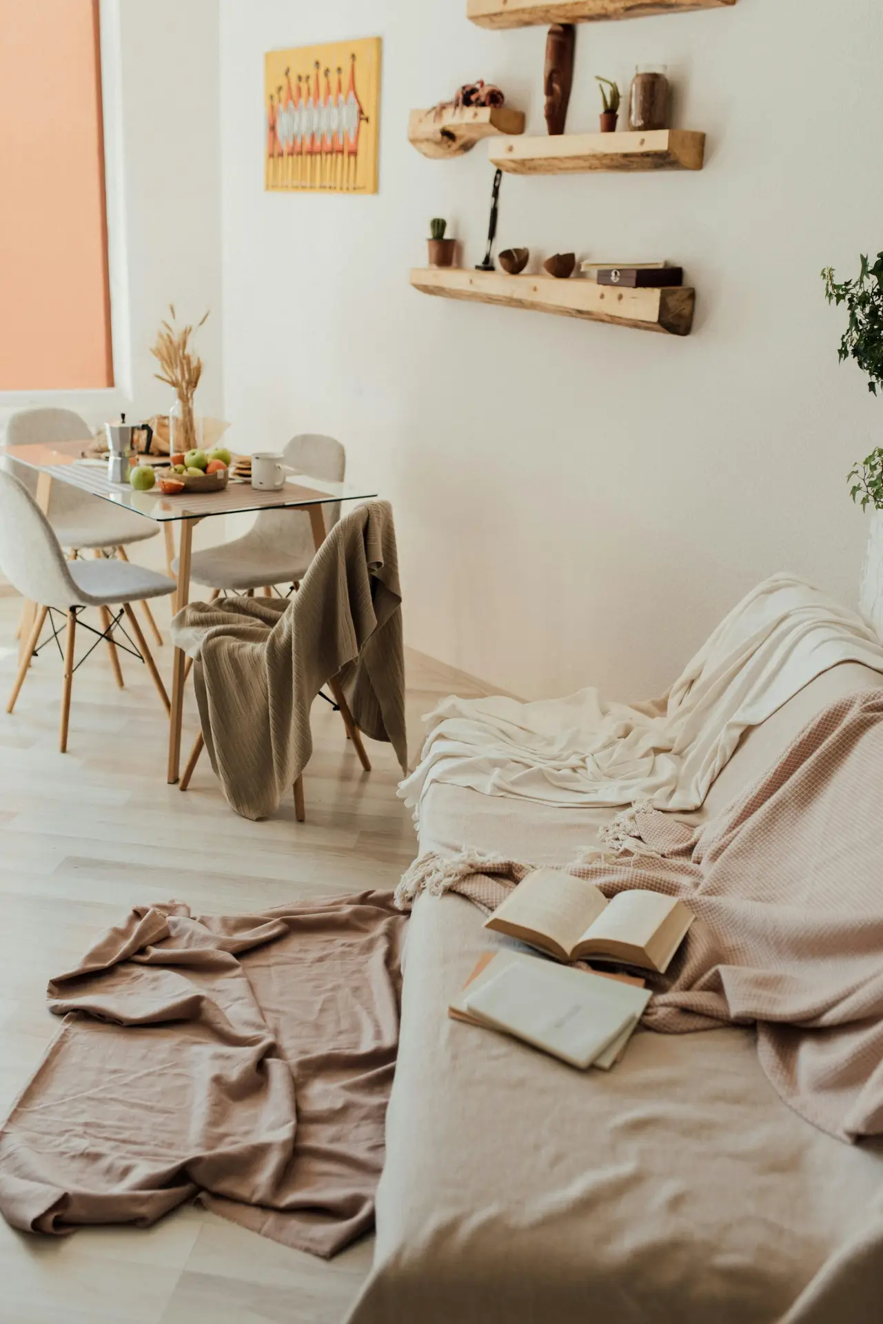 A warm and inviting open-plan living area demonstrating cozy small apartment decorating ideas. In the foreground, a beige sofa is draped with several soft blankets, with an open book resting on the cushions. The background features a glass dining table set for a meal with light grey chairs. On the wall above the sofa, several rustic wooden floating shelves are arranged at varying heights, displaying small potted succulents, decorative bowls, and an African-style mask. A vibrant yellow piece of art adds a pop of color to the neutral-toned walls and light wood flooring.