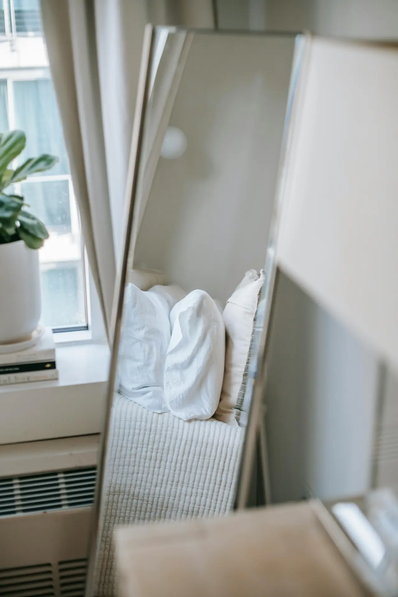 A brightly lit bedroom corner showcasing stylish small apartment decorating ideas. A tall, arched freestanding mirror with a thin black frame leans against a light-colored wall, reflecting the surrounding space including a woven basket and a plant. The floor is made of large, square grey tiles. To the right, a queen-sized bed is made with light grey bedding, a chunky beige knit throw, and an assortment of pillows with varying textures and patterns. A minimalist, multi-arm black chandelier hangs from the white ceiling, and a large potted fiddle-leaf fig tree stands prominently in the foreground next to the bed, adding organic greenery.