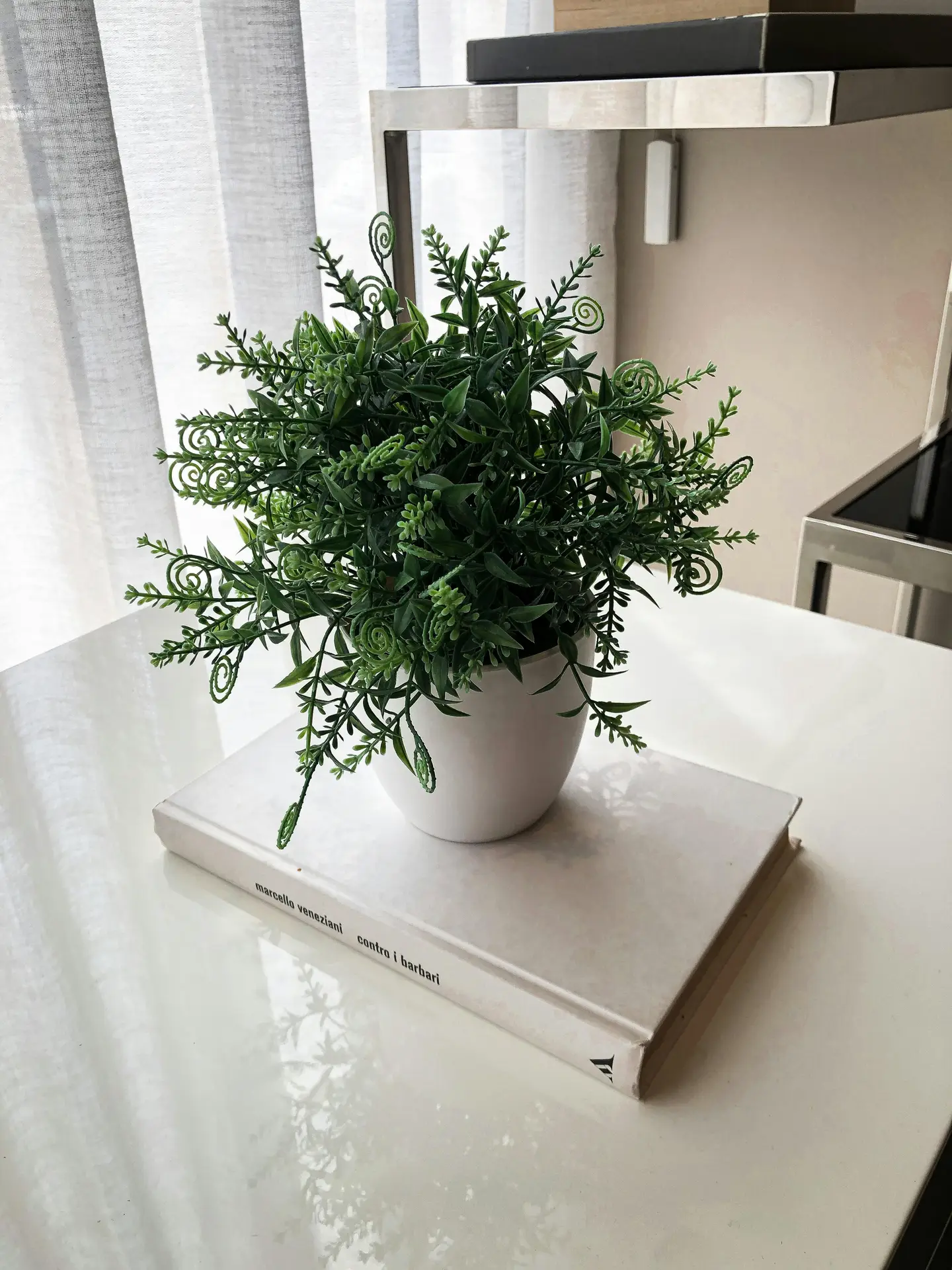 A close-up shot of a polished white surface illustrating how to keep decor simple for effective small apartment decorating ideas. A small, vibrant green artificial plant in a minimalist white ceramic pot rests atop a single large hardcover book with a white spine. The arrangement is positioned near a window with soft, sheer grey curtains that diffuse the natural light, while a sleek chrome furniture frame is partially visible in the background, emphasizing a clean and uncluttered aesthetic.