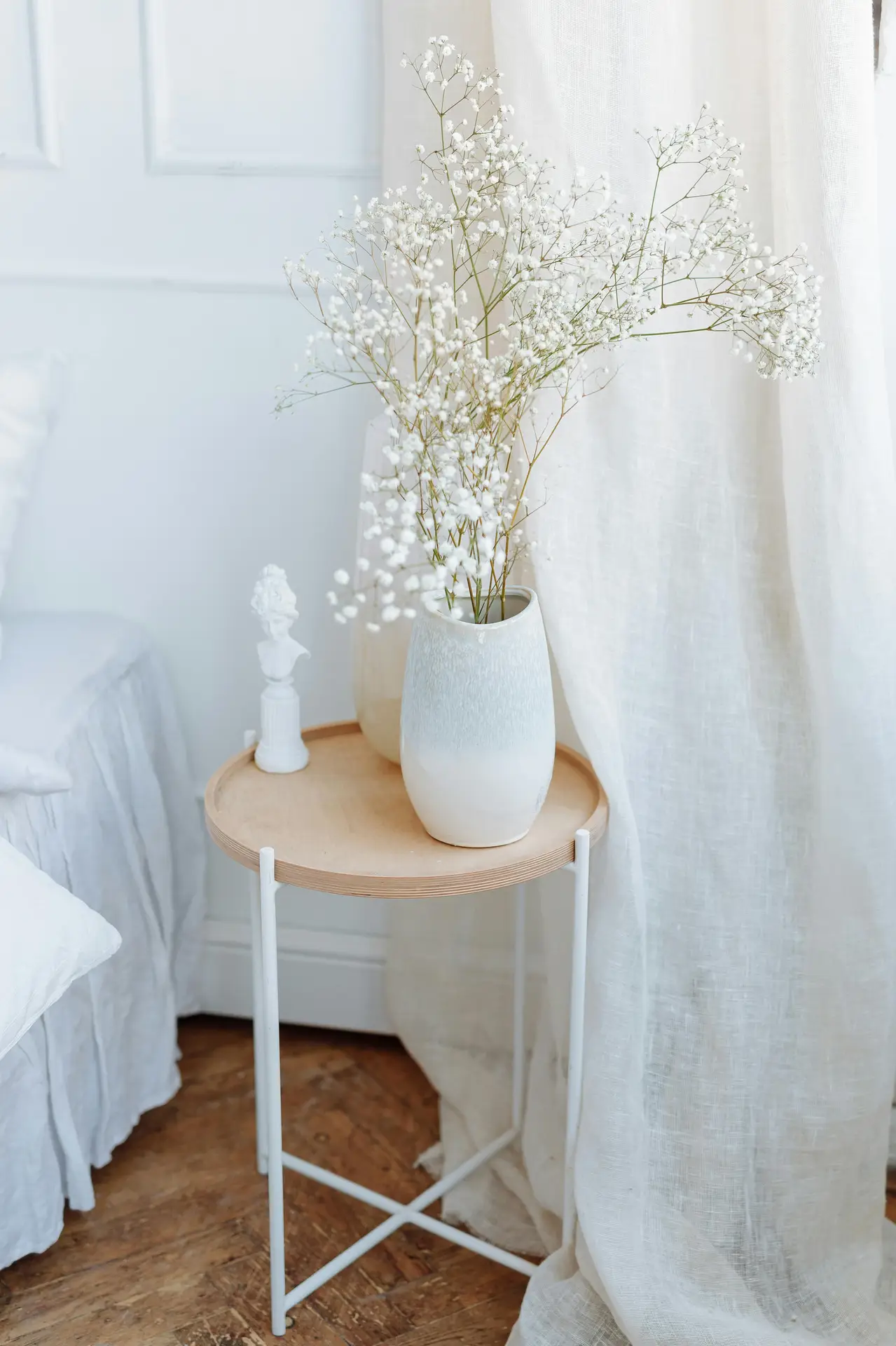 A dreamlike bedroom corner featuring small apartment decorating ideas with a focus on airy textures. A minimalist round side table made of light wood and white metal legs holds a ceramic vase filled with delicate light wood and white flowers, specifically baby's breath. Beside the vase sits a small white classical bust sculpture. The scene is softened by flowing white linen curtains and a white bed skirt, all resting on a rustic hardwood floor, creating a bright and peaceful atmosphere.