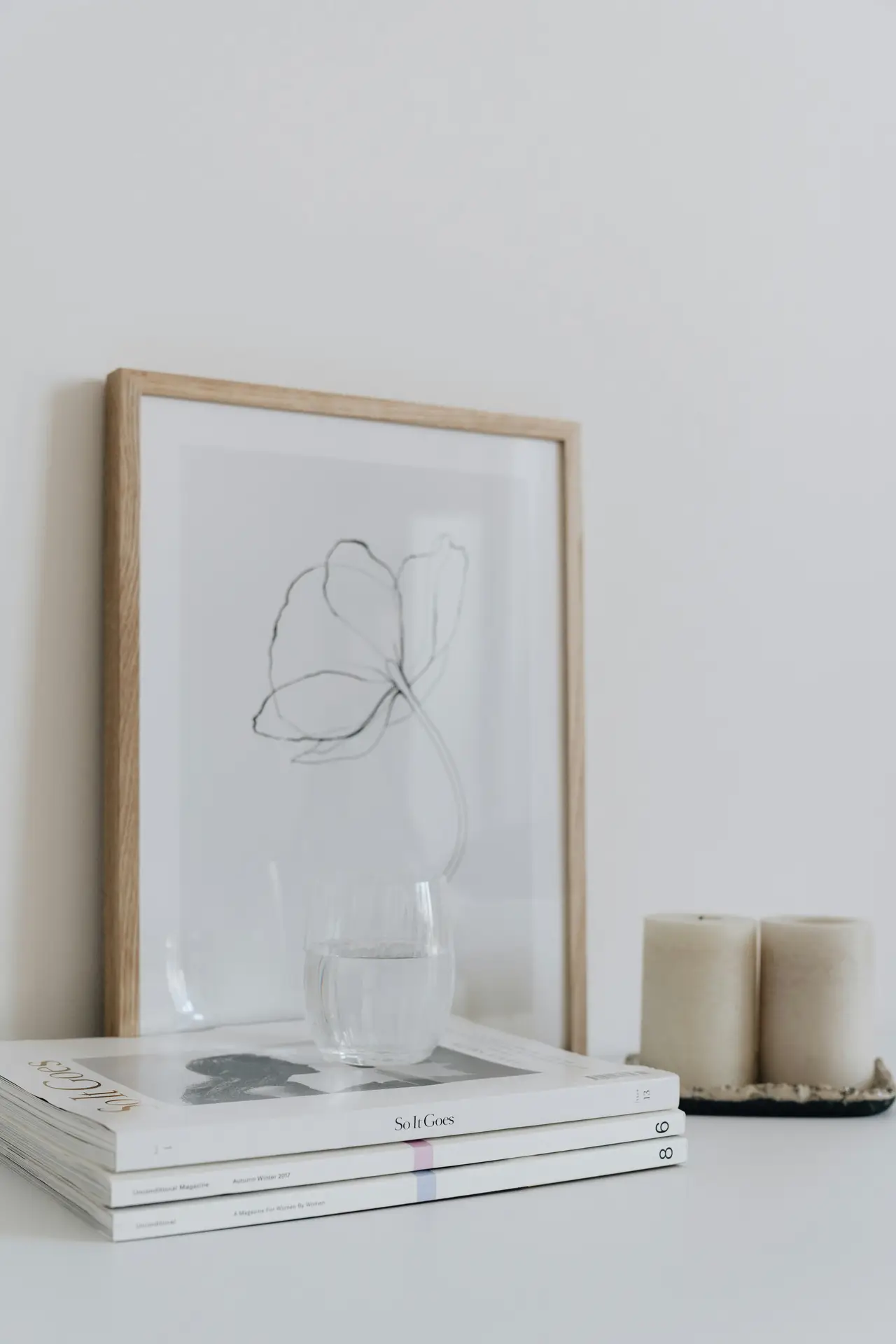 A minimalist and aesthetic vignette showcasing small apartment decorating ideas. The scene features a stack of magazines, candles and picture in frame arranged on a white surface against a plain wall. A light wood frame holds a delicate line drawing of a flower, partially obscured by a clear glass of water. Next to the magazines, two thick cream-colored pillar candles sit on a small dark tray, creating a calm and sophisticated atmosphere.