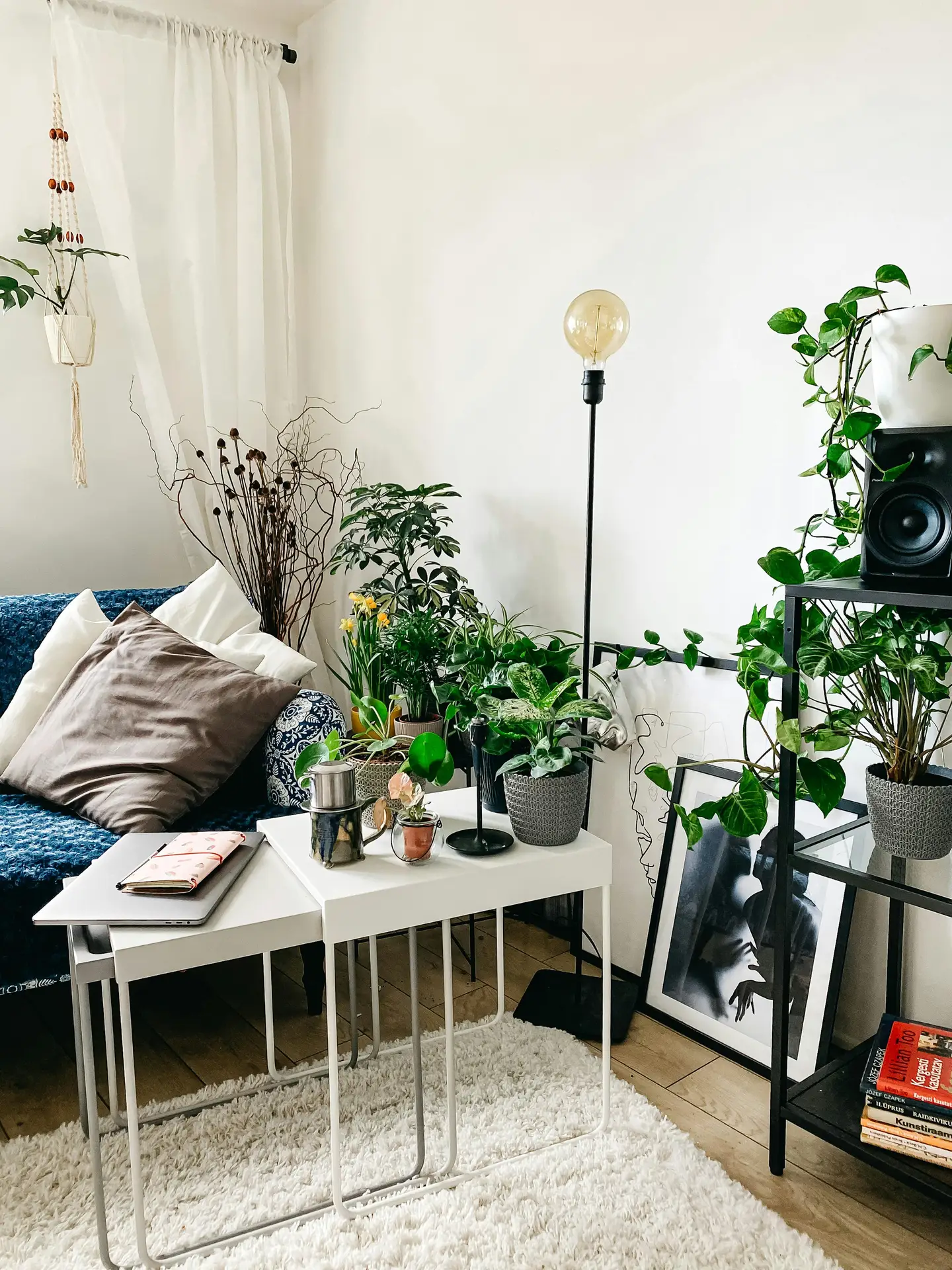 An eclectically decorated living room filled with greenery, demonstrating smart small apartment decorating ideas. A set of white nesting tables serves as a versatile coffee table, holding a laptop, a notebook, and various small potted plants. The cozy space features a blue patterned sofa with large brown and white pillows, a shaggy white rug on wooden floors, and a black minimalist floor lamp with an oversized bulb. A black shelving unit on the right displays more trailing plants and books, while framed black-and-white art leans against the wall, contributing to the layered, botanical aesthetic.