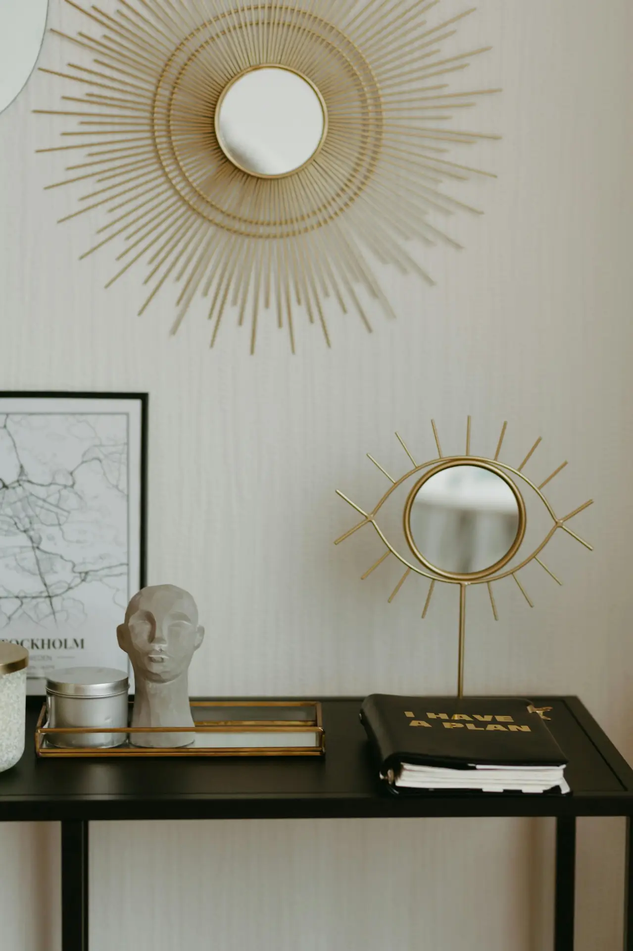 A chic and curated display that uses recurring elements to create a cohesive look, perfect for small apartment decorating ideas. On a black console table, a small eye-shaped gold mirror on a stand is paired with a matching sunburst mirror mounted on the wall above. The arrangement includes a grey bust sculpture on a gold-rimmed glass tray, a silver tin, and a black notebook with the words 