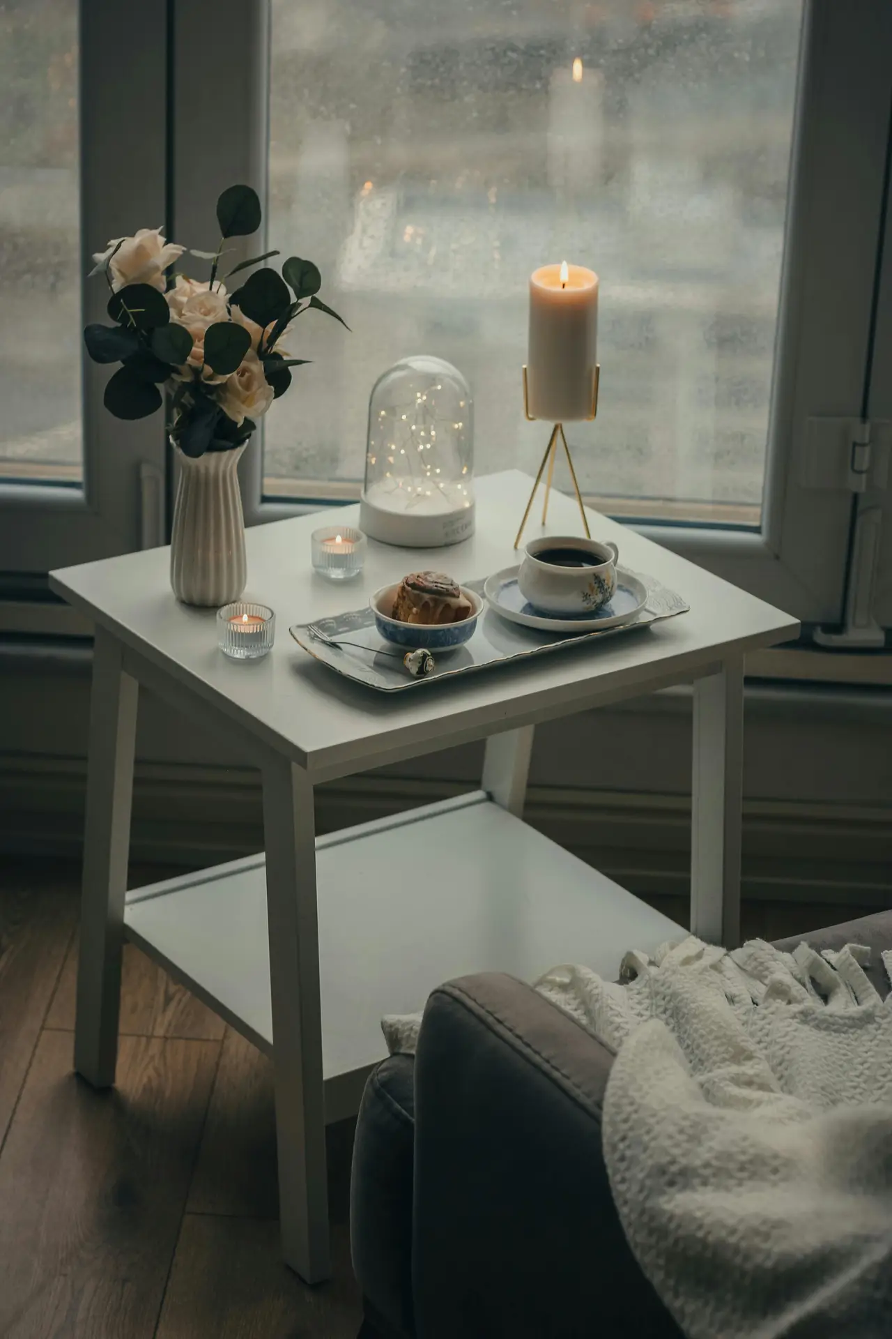 A cozy afternoon setting that offers charming small apartment decorating ideas. A white two-tiered side table is placed next to a grey armchair draped with a chunky white knit throw. On the table, a decorative tray holds a cup of coffee and a cinnamon roll, surrounded by a white vase of roses, a lit pillar candle on a gold stand, and a glass cloche filled with fairy lights. The scene is set against a large window with a view of a rainy day, creating a warm and inviting indoor atmosphere.