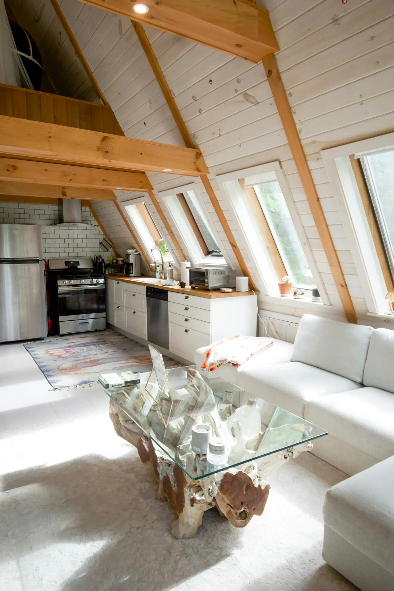A bright, A-frame attic living space illustrating bold small apartment decorating ideas. The open floor plan features a white kitchen with wood countertops and stainless steel appliances alongside a cozy lounge area with a plush white sofa. A unique driftwood coffee table with a glass top serves as a natural statement piece in the center of the room. Sunlight pours through a series of large skylights in the slanted white wood-paneled ceiling, illuminating the neutral rug and creating an airy, expansive feel.
