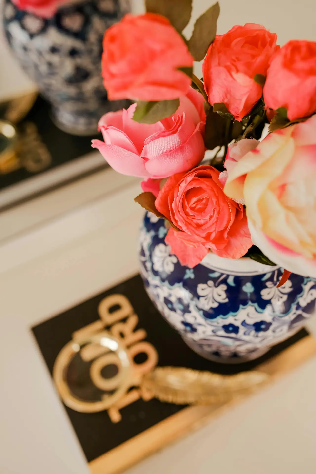 A vibrant and charming tablescape vignette illustrating summer decor ideas. At the center, a clear glass vase holds a lush bouquet of coral ranunculus, pink roses, and eucalyptus. It sits on a round wooden tray next to a woven round placemat, which is layered with a scalloped-edge white plate and a light blue linen napkin. Gold-handled cutlery and a matching blue ceramic salt cellar with a spoon complete the place setting on a weathered wood table. Natural light streams from a window to the right, illuminating a patterned blue cushion on a wicker chair and a woven basket in the blurred background.