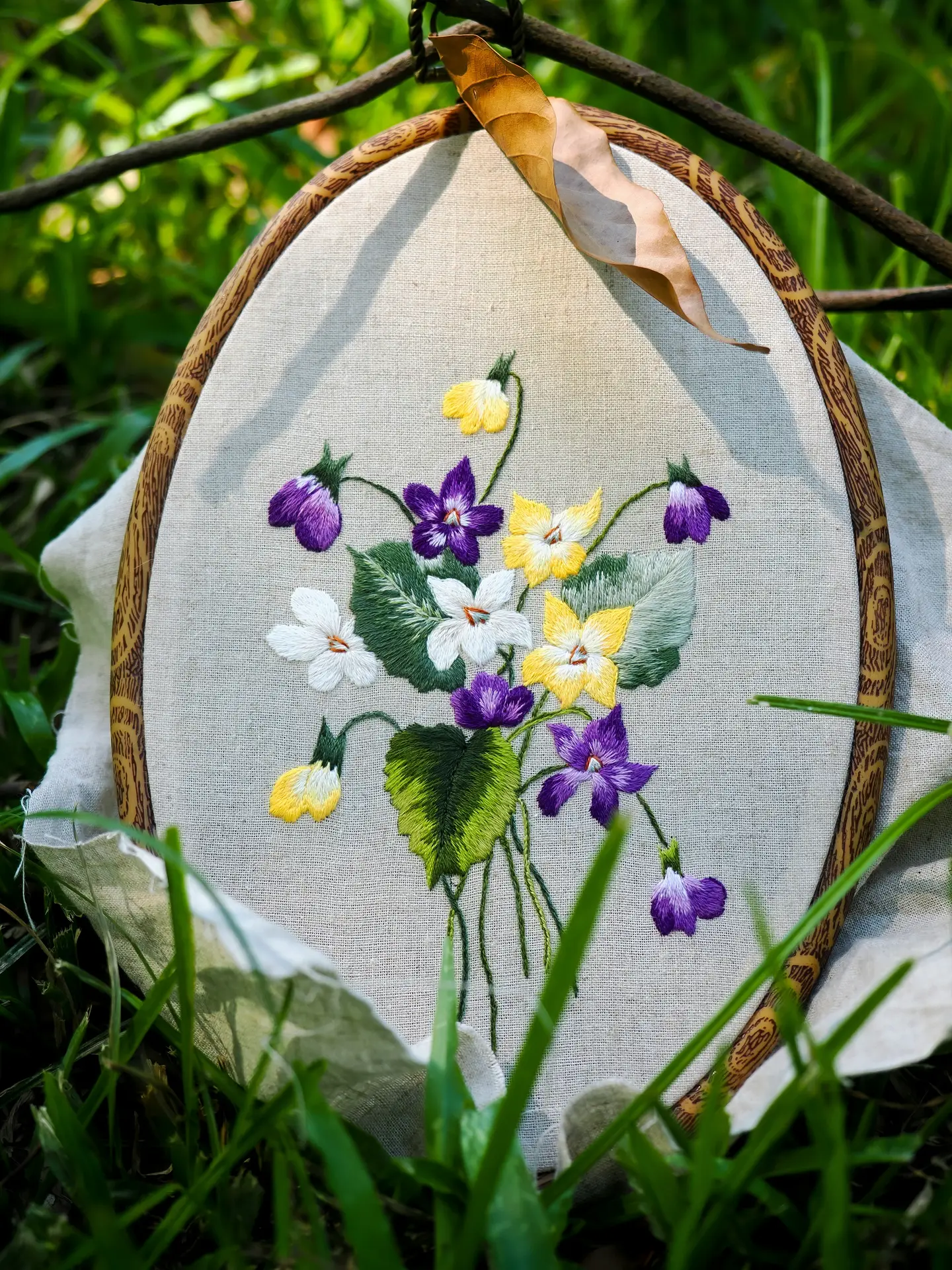 A delicate and handcrafted piece that serves as inspiration for summer decor ideas. The image shows an oval wooden embroidery hoop framing a beautiful bouquet of embroidered flowers on light linen fabric. The design features a mix of purple violets, white blossoms, and yellow primroses with detailed green leaves, all stitched with vibrant thread. The hoop is leaning against natural green grass in soft sunlight, with a single dried leaf resting at the top, emphasizing a fresh and botanical summer aesthetic.