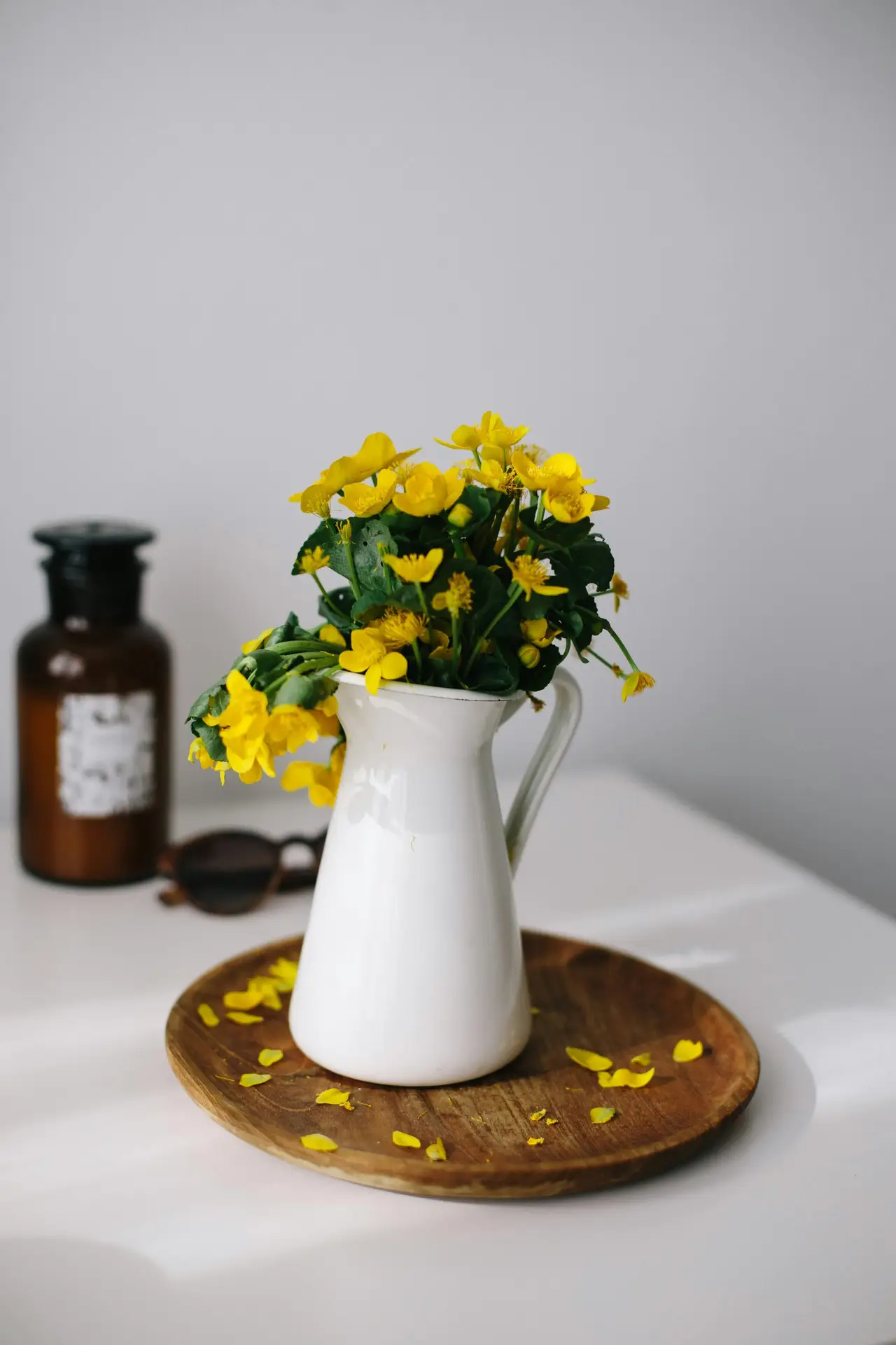 A bright and cheerful display offering simple summer decor ideas. The image features a vibrant bunch of yellow wildflowers arranged as flowers in milk can, with the white enamel pitcher sitting atop a round wooden tray. Scattered yellow petals rest on the wood surface, adding a natural touch. In the soft-focus background, a brown apothecary-style glass bottle and a pair of sunglasses sit on the white tabletop, all bathed in clear, natural light.