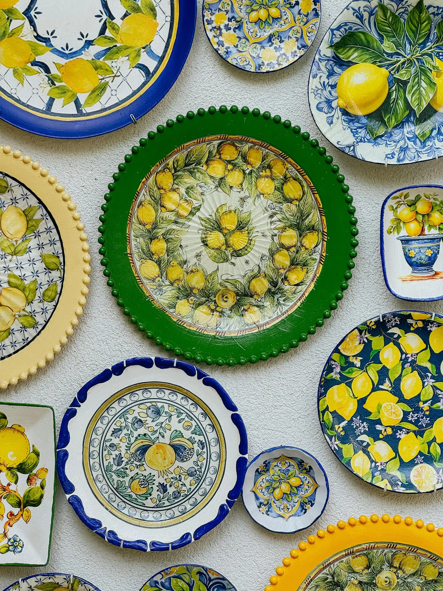 A close-up shot of a white kitchen counter where a large, clear glass bowl is generously filled with bright yellow lemons and fresh green leaves, serving as a simple and cheerful summer decor idea. Behind the bowl, which sits on a wooden cutting board with a white linen towel, a glass pitcher holds water infused with lemon slices and mint. The background includes white subway tiles, a wooden-framed botanical print of lemons, and a white crock with wooden utensils, all illuminated by soft natural light from a window looking onto a green garden.