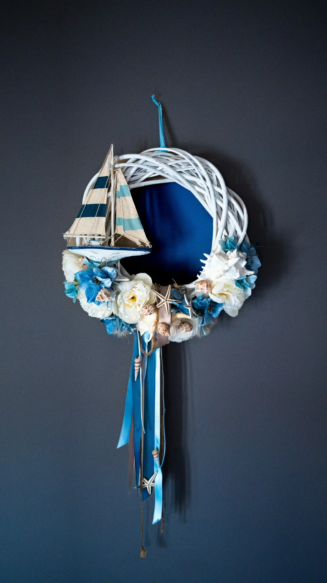 A decorative maritime wreath made of white woven branches hanging against a dark grey wall, showcasing nautical summer decor ideas. The wreath is embellished with white and blue fabric flowers, assorted seashells, and small starfishes. A small wooden sailboat with blue-and-white striped sails is tucked into the left side. Long blue and light blue satin ribbons, some adorned with tiny shells and a starfish, hang elegantly from the bottom of the arrangement.