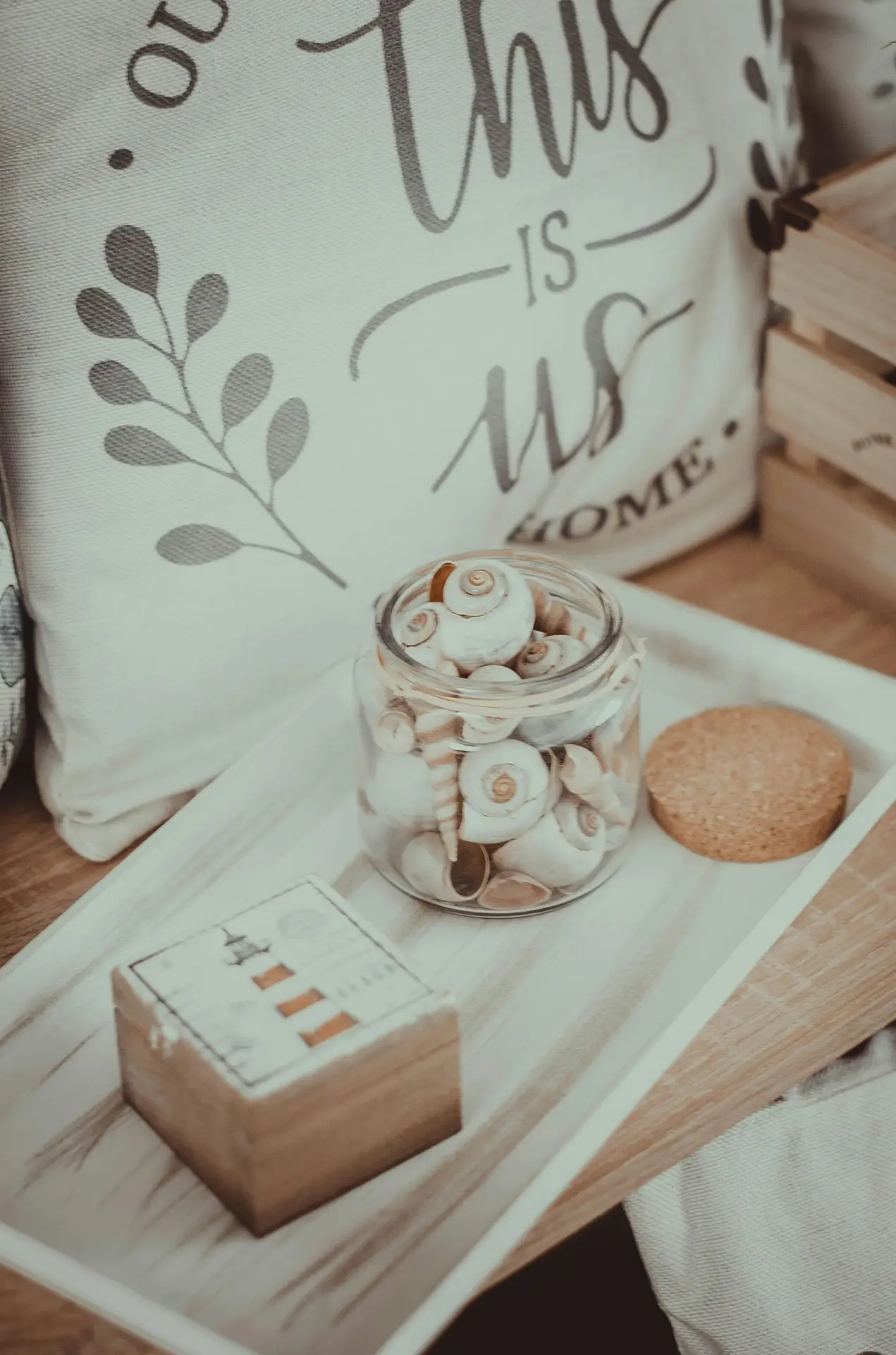 A charming close-up photograph of a windowsill display that incorporates fresh summer decor ideas. At the center, a blue and white patterned ceramic pot, featuring a wave-like design, holds a small, vibrant green fern. A cluster of small, varied seashells is nestled at the base of the plant. Hanging from the pot by a thin string are two small, white-painted wooden fish figurines with blue stripe maritime details. The display sits on a distressed white-painted wooden sill, with natural daylight filtering through the glass pane, casting soft shadows against the window frame and a rustic white wall, creating a breezy, coastal feeling.