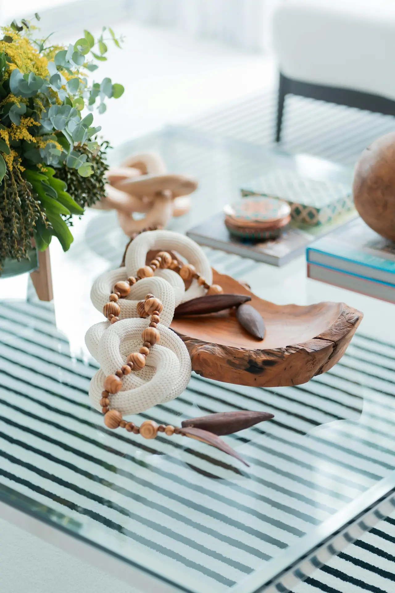 A top-down perspective of a glass coffee table styled with organic summer decor ideas and tactile natural details. A live-edge wooden bowl serves as a centerpiece, holding dark seed pods and draped with a string of polished wooden beads and thick, white knotted rope links. In the background, a lush floral arrangement of yellow blossoms and eucalyptus leaves sits beside a stack of books and a small decorative tin. The table rests over a bold black-and-white striped rug, creating a sophisticated and breezy interior aesthetic.