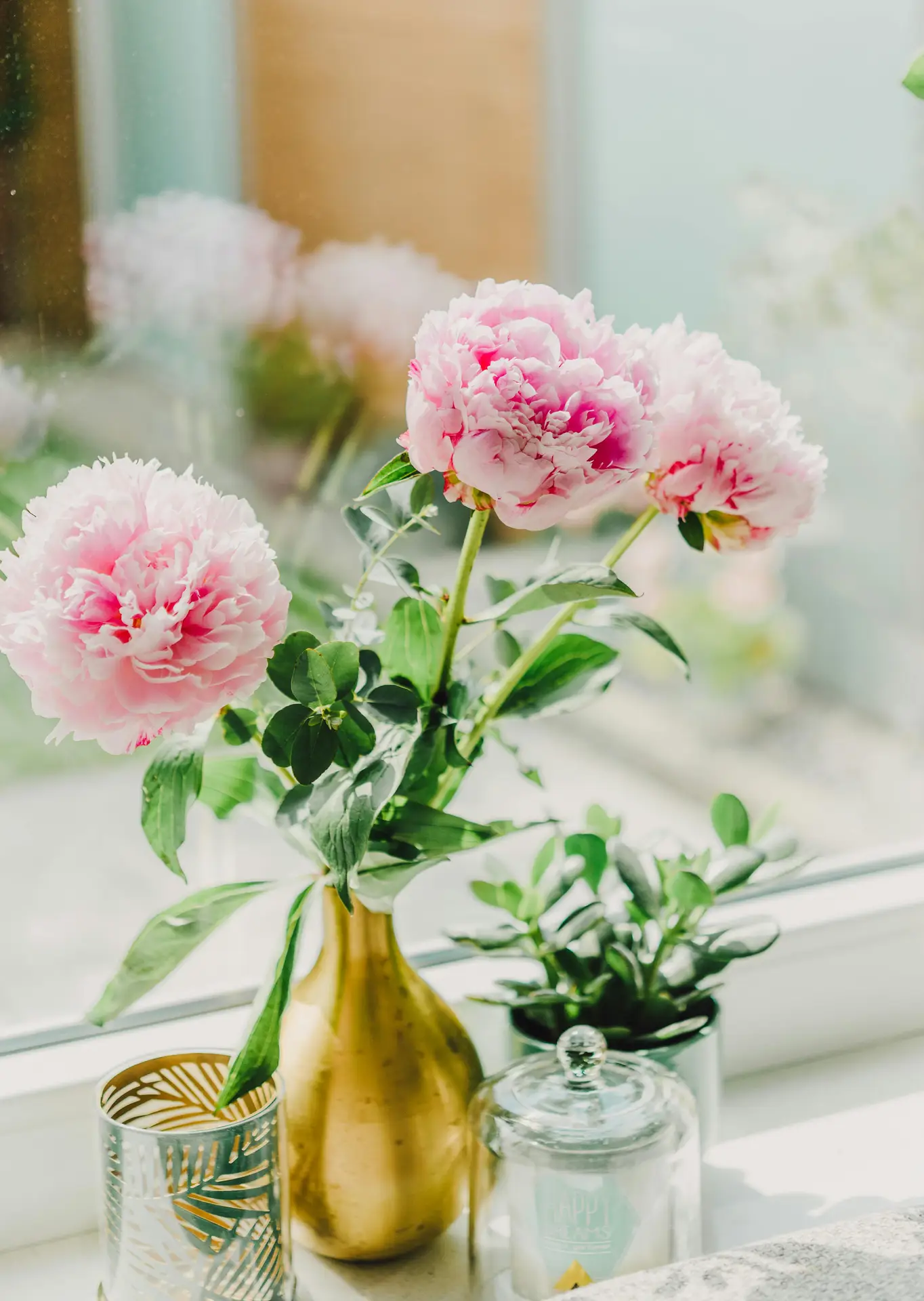 A bright windowsill arrangement featuring elegant summer decor ideas with a striking pink and gold theme. A slender gold vase holds three large, lush pink peonies and sprigs of eucalyptus, catching the soft afternoon light. Next to the bouquet sits a small green succulent in a pot, a patterned gold tea light holder, and a glass candle jar with a lid. The soft-focus background shows a view through the window, enhancing the airy and feminine feel of the decorative display.