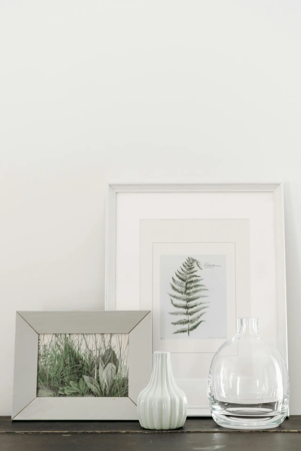 A minimalist and calming display on a dark wooden surface, perfect for summer decor ideas with serene aesthetics. The arrangement consists of a large white frame featuring a botanical fern print and a smaller grey frame showcasing a detailed illustration of green meadow grass. In front of the frames, a small, pale mint-colored fluted ceramic vase and a round, clear glass decanter add a clean, airy touch against the simple white wall, creating a peaceful and refreshing seasonal atmosphere.