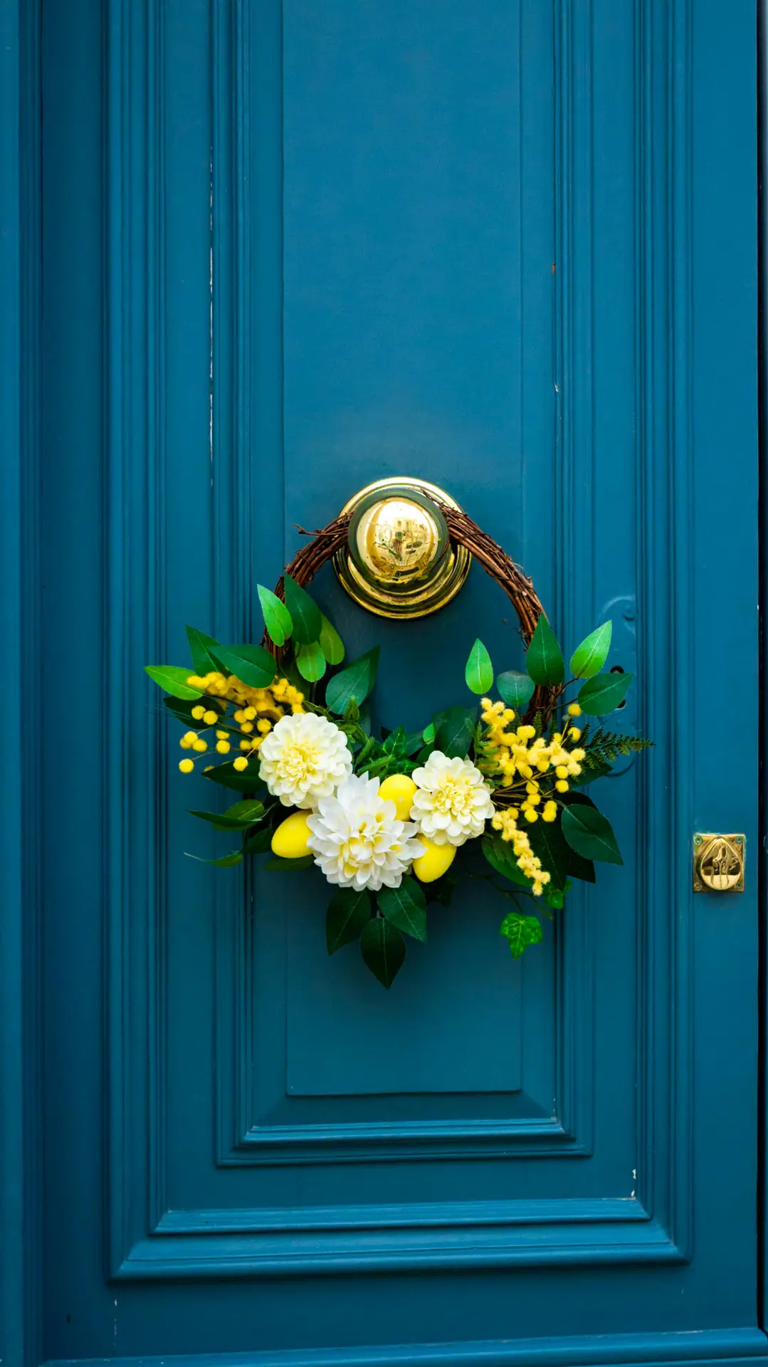 A vibrant teal-colored wooden door serves as the backdrop for beautiful summer decor ideas. Hanging from a polished gold door knob is a rustic grapevine summer flower wreath, decorated with white dahlias, bright yellow mimosa blossoms, and lush green leaves. The composition is balanced and elegant, featuring a pop of yellow from small decorative lemons nestled among the flowers, creating a welcoming and sunny entryway.