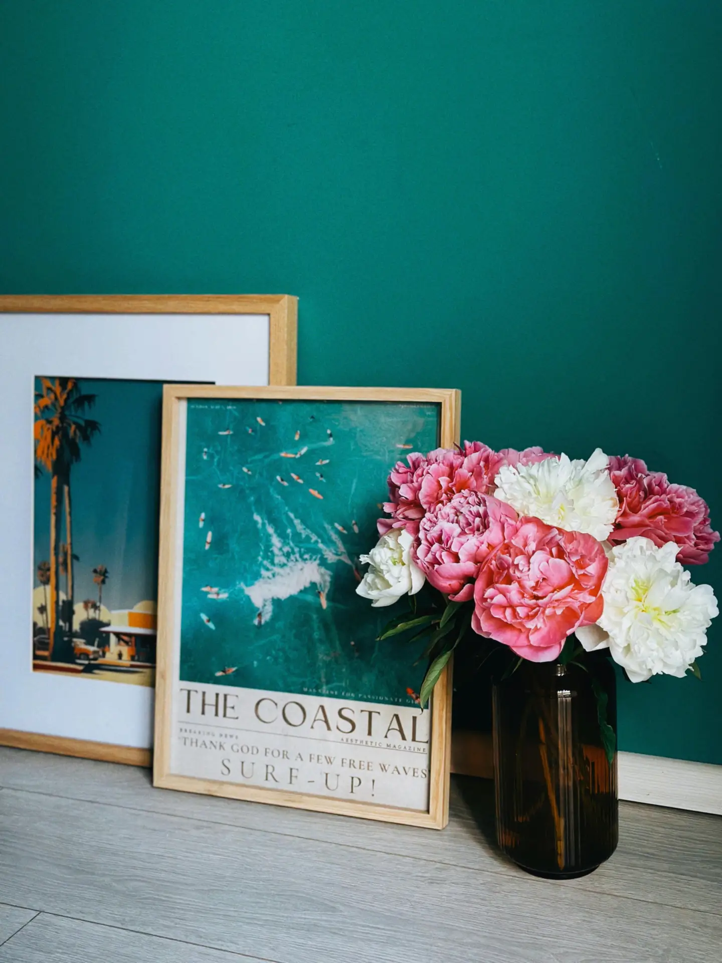 A cheerful and vibrant living room display showcasing inspiring summer decor ideas. Three colorful summer prints in frames on the floor are leaning against a clean white wall with horizontal paneling. The central print is larger, featuring an abstract face with botanical motifs and text like 