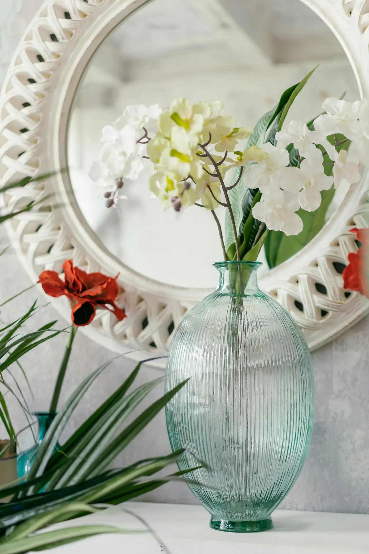 A beautiful close-up photograph capturing elegant summer decor ideas on a rustic white-painted wooden windowsill. A vibrant white orchid plant with several blossoms and buds in full bloom is featured in a white ceramic pot with a textured pattern. Next to the orchid, there is a round, clear glass jar filled with various natural items like sand, small seashells, and smooth pebbles, topped with a jute rope. To the right, a white wooden-framed botanical print of a fern leaf leans against the windowpane. Sunlight filters through the glass, illuminating the scene and creating a bright, breezy coastal atmosphere. The window overlooks a blurred garden with green foliage and a hint of a white fence.