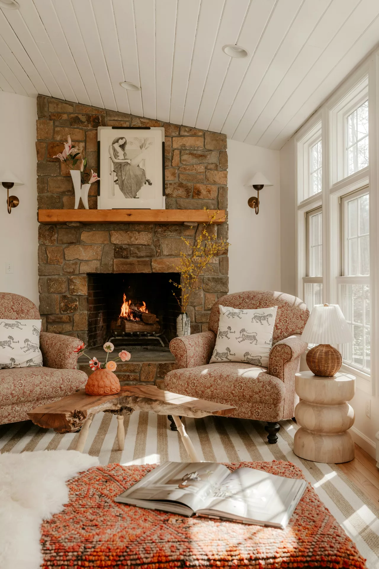 A cozy and warm living room scene featuring inspiring summer mantel ideas. Above the stone fireplace, a wooden mantel displays a Big Photo Frame & Flowers, with a black-and-white art print of a seated woman and a sculptural white vase holding pink lilies. Two floral-patterned armchairs with zebra-print cushions flank the hearth, where a fire is burning. In the foreground, a live-edge wooden coffee table holds a small orange vase with delicate blooms, while a light-filled window and a unique stacked wooden side table with a wicker lamp complete the comfortable, rustic interior.