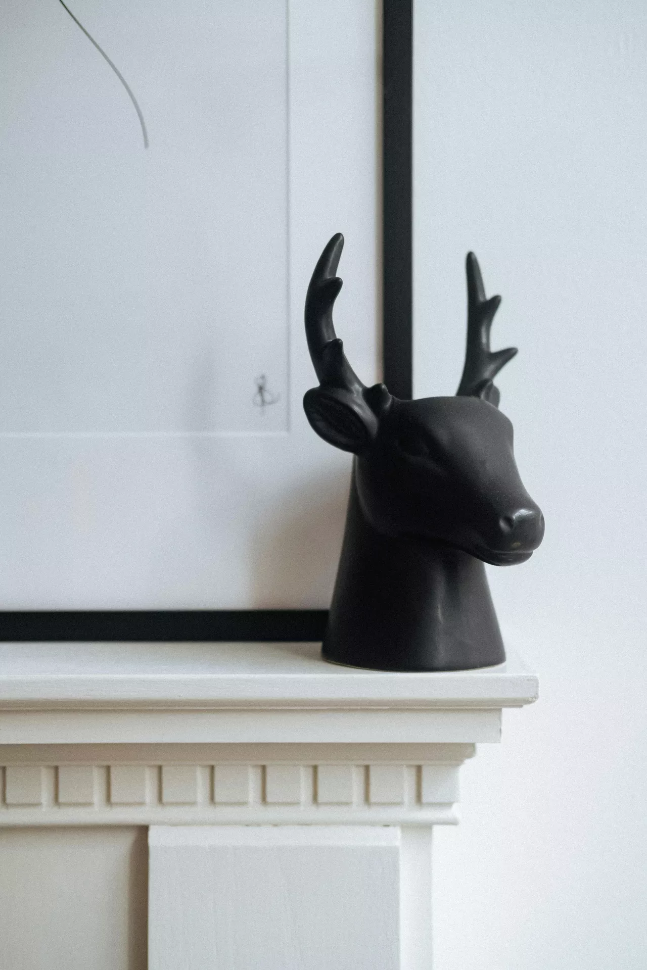 A close-up shot of a rustic wooden mantelpiece adorned with summer mantel ideas that incorporate black accents. Leaning against a white shiplap wall is a large, vertical art print featuring stylized black and beige botanical illustrations in a thin black frame. To its left sits a cylindrical vase in matte black, holding several leafy green olive branches and small cluster of small yellow wildflowers. Two small white pillar candles rest on a small black ceramic tray next to the vase. On the right, a round woven mirror with a jute frame adds a natural texture, accompanied by two stack of black-spined books. A grey linen basket holds extra branches and textiles below.