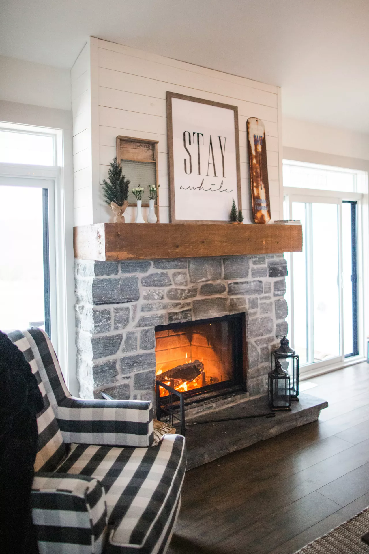 A cozy and inviting stone fireplace serves as a central focal point for these rustic summer mantel ideas with boho vibes. A thick wooden mantel beam sits atop the grey stonework, displaying a large framed sign that reads 