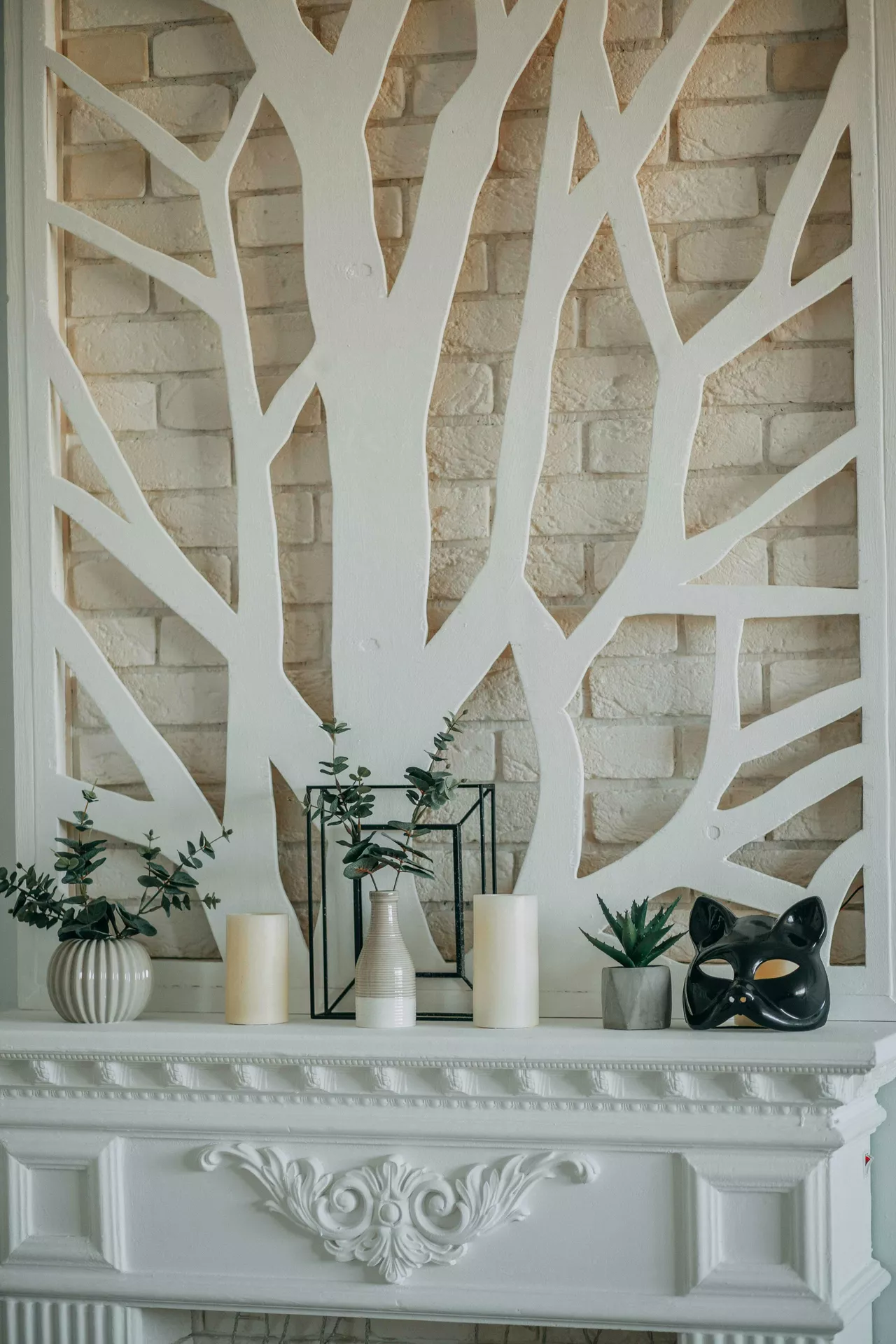A sophisticated white mantelpiece decorated with modern summer mantel ideas featuring a balanced mix of candles and greenery. Positioned against a beige brick wall with a large white decorative tree-branch cutout, the display includes various potted plants such as eucalyptus in a ribbed grey vase and a small succulent in a concrete pot. Two tall, cream-colored pillar candles flank a central geometric black wire frame that surrounds a slender striped bud vase. On the far right, a glossy black cat mask adds a unique, artistic touch to the bright and airy arrangement.