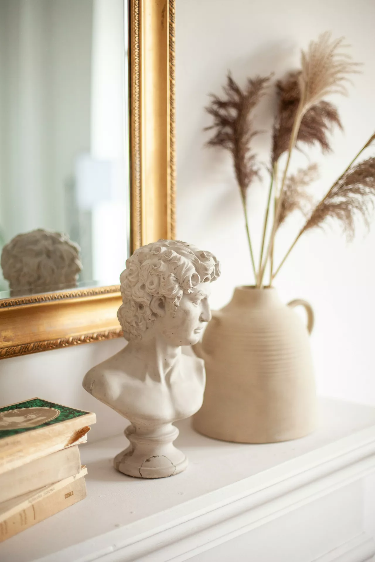 An elegant and timeless arrangement on a white mantelpiece, offering sophisticated summer mantel ideas for a classic interior. A classical plaster bust of a young man with curly hair serves as the focal point, positioned next to a large gold-framed mirror that reflects the bright room. Behind the bust stands a beige ceramic jug with two handles, filled with tall, feathery dried pampas grass. On the left, a small stack of vintage books with worn edges and a green cover adds a scholarly touch to the bright, airy scene.