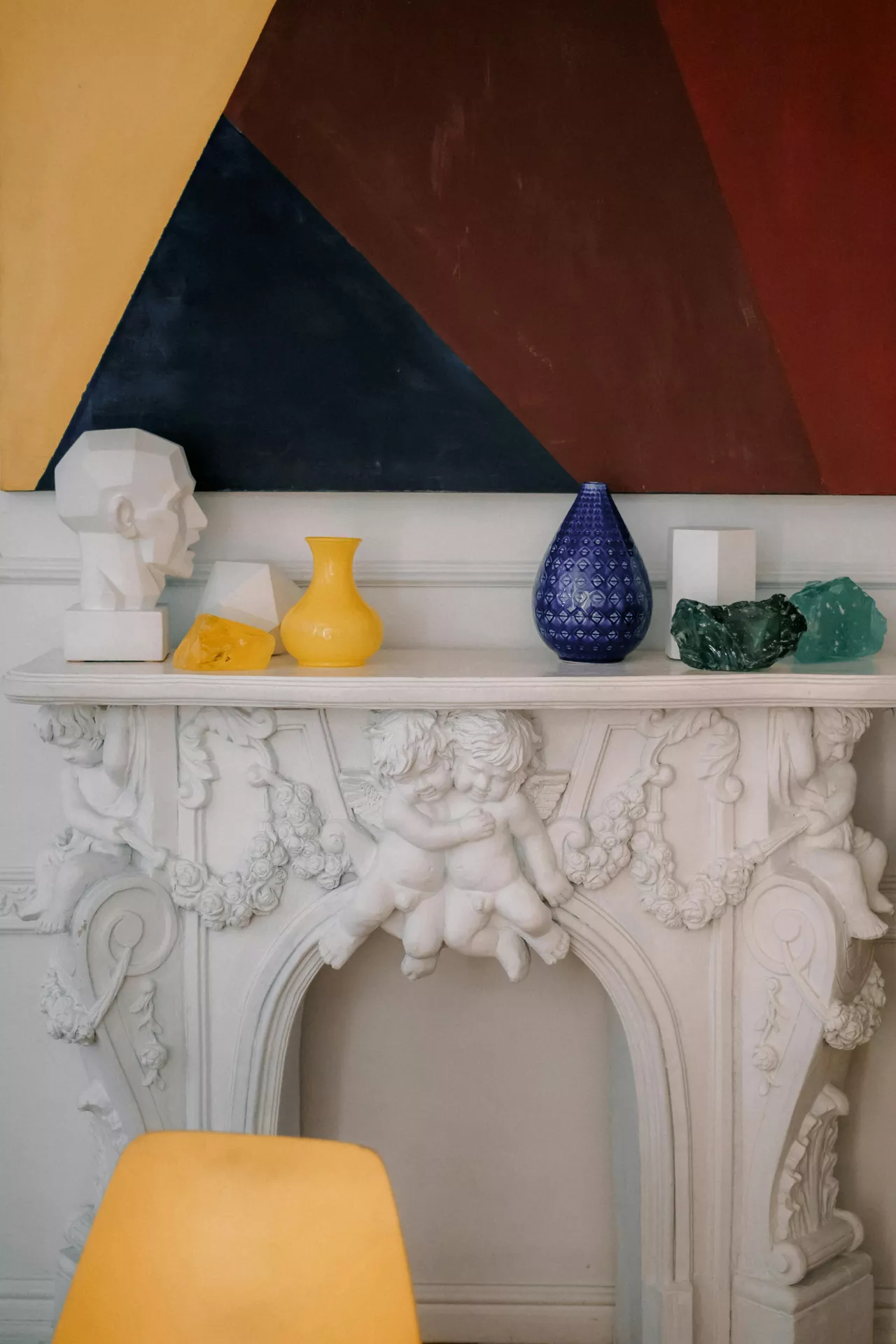 
A striking white ornate fireplace with elaborate cherub carvings is used as a base for modern summer mantel ideas. The display is vibrant, featuring a colorful and geometric theme with a low-poly white bust, a bright yellow vase, and a textured royal blue teardrop-shaped vessel. Accompanying these pieces are several glass-like decorative rocks in translucent amber and emerald green. The entire arrangement is set against a large, bold abstract painting on the wall with sharp diagonal sections of navy blue, deep red, and yellow, creating a playful contrast between classical architecture and contemporary design.