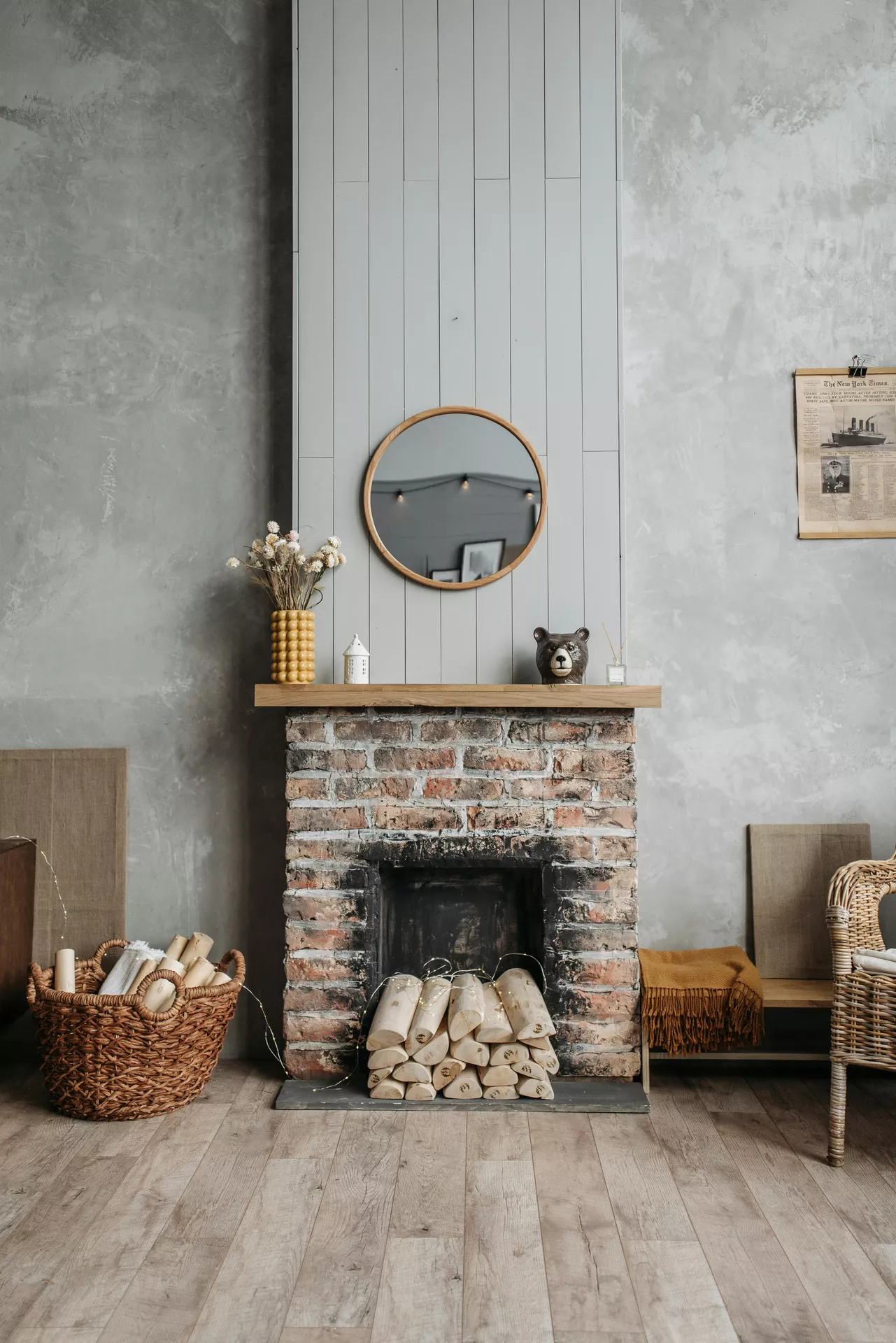 A rustic brick fireplace with a light wood mantel showcases charming summer mantel ideas that evoke cozy cabin vibes. On the mantel sits a textured yellow vase with dried white flowers, a small white house-shaped lantern, and a decorative brown bear head. A simple round mirror with a wooden frame hangs above on a grey paneled wall. In the fireplace opening, a stack of birch logs is decorated with delicate fairy lights. The surrounding space features a large woven basket filled with logs, a wicker armchair, and a mustard-yellow throw blanket, all set against a textured concrete wall and light wood flooring.