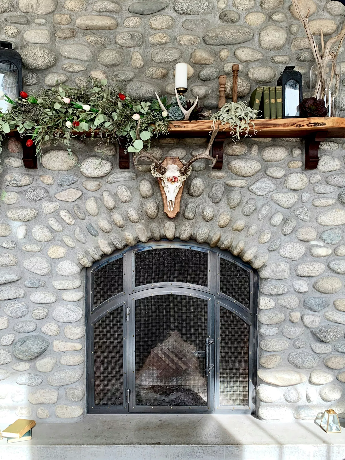 A large, rustic fieldstone fireplace serves as a backdrop for eclectic summer mantel ideas. The live-edge wooden mantel is decorated with a lush garland of eucalyptus and pink-tipped flowers on the left, while the right side features deer antlers, flowers & vintage accessoires, including a row of green antique books, wooden candlesticks, and a dark lantern. Centered just below the shelf on the stones is a mounted deer skull adorned with a delicate floral wreath. The scene is completed by a curved black metal fireplace screen and soft natural light hitting the hearth.