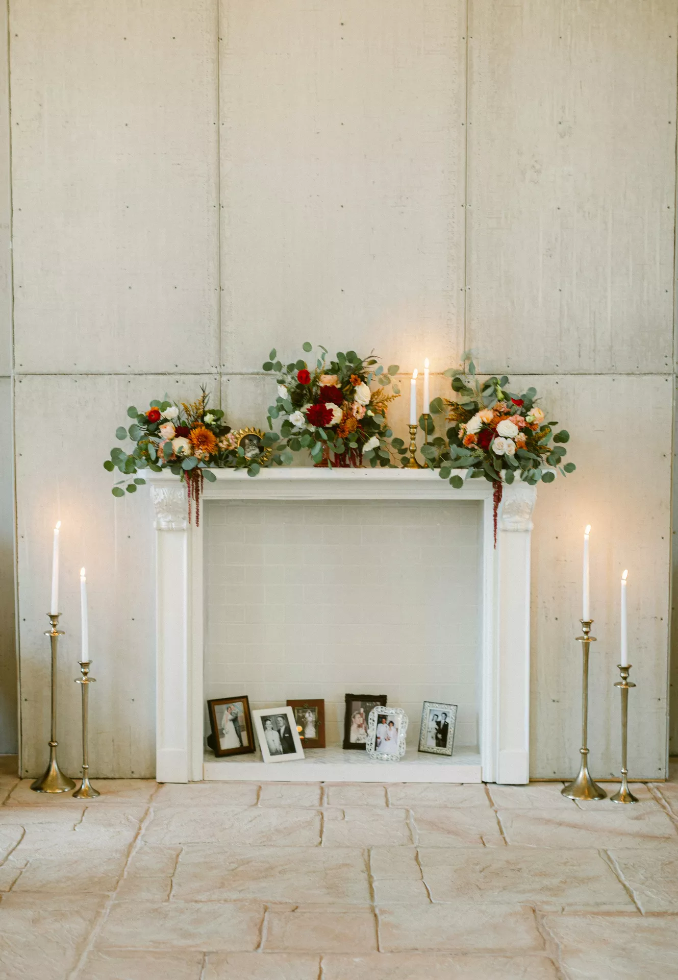A minimalist white fireplace mantel set against a large paneled wall, showcasing elegant summer mantel ideas. The top of the mantel is adorned with three lush floral arrangements featuring eucalyptus leaves and deep red and orange blossoms, interspersed with slender white lit candles in gold holders. Within the fireplace opening, a sentimental display of various Flowers, Photo Frames & Candles continues, with several black-and-white family photographs in ornate frames resting on the hearth. Tall, brass floor candlesticks with glowing tapers flank the fireplace on the stone-tiled floor, creating a warm and reverent atmosphere.