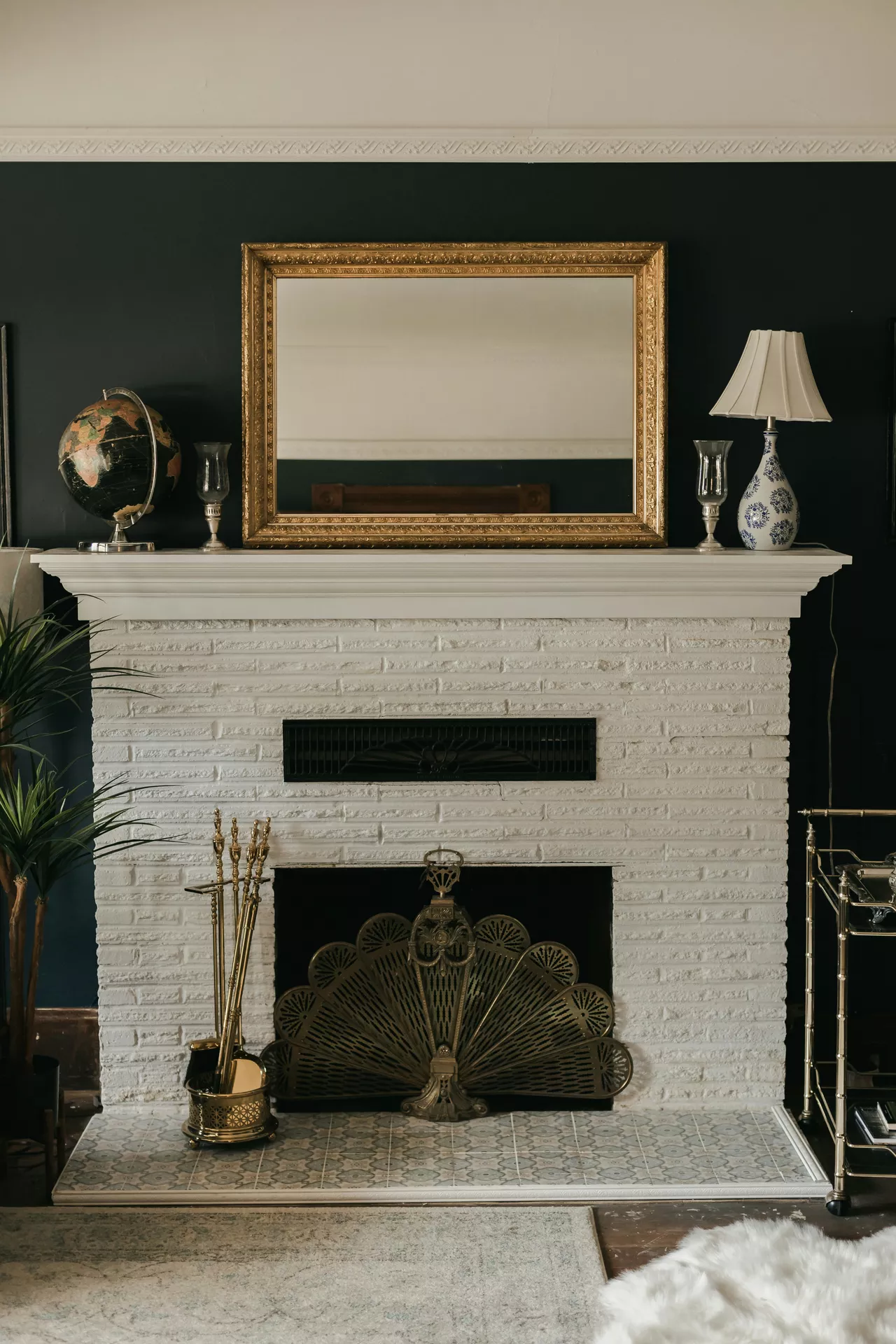 A classic white brick fireplace is the center of these traditional summer mantel ideas, set against a dark moody wall. Resting on the mantel shelf is a large, ornate horizontal mirror with a thick gilded frame that reflects the room's light. The mantel is further decorated with a vintage globe on the left and a blue-and-white patterned lamp with a pleated shade on the right, flanked by two slender glass candle holders. An intricate brass fan-shaped fire screen covers the hearth, accompanied by matching brass fireplace tools on a patterned tile base, creating a sophisticated and timeless aesthetic.