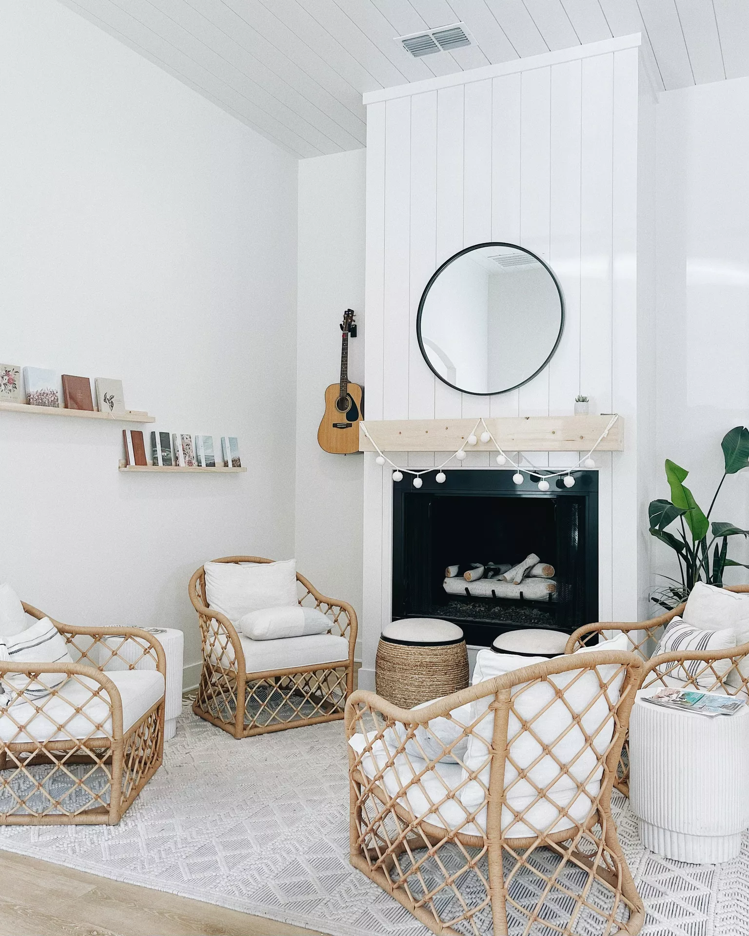 A bright and airy living room featuring minimalist summer mantel ideas. A light wood mantel is decorated with a delicate white pom-pom garland, while a simple round black mirror hangs above on a white shiplap wall. Surrounding the fireplace are four rattan armchairs with white cushions, a woven jute ottoman, and a large green plant, creating a relaxed coastal vibe. Although lanterns are not physically present on the shelf, the overall bright aesthetic provides a perfect backdrop for such glowing accents to enhance the seasonal atmosphere.