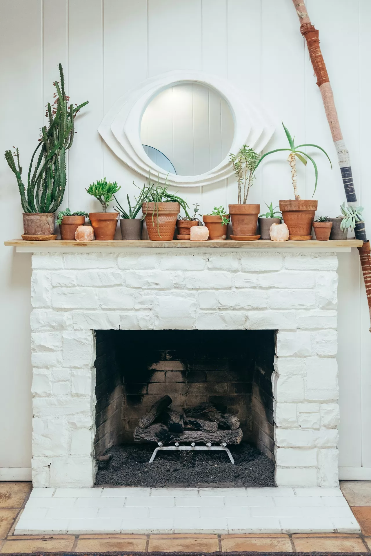 A vibrant white-painted stone fireplace featuring a thin wooden mantel shelf packed with diverse summer mantel ideas. The display showcases lots of plants in various terracotta and clay pots, including tall cacti, sprawling succulents, and several aloe vera varieties. Nestled between the pots are small pink Himalayan salt chunks, adding a natural glow to the arrangement. Above the greenery hangs a unique, eye-shaped white mirror on a shiplap wall, while a decorative woven walking stick leans against the side. The scene is grounded by a dark hearth with logs ready for a cool summer evening.