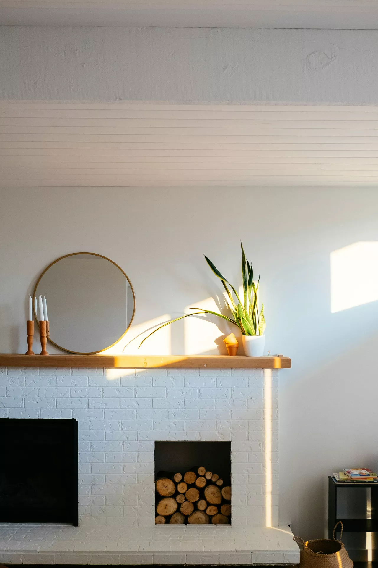 A clean white brick fireplace bathed in warm afternoon sunlight, featuring summer mantel ideas with a distinct minimalist style. The simple wooden mantel shelf holds a gold-rimmed circular mirror, three white taper candles in light wood holders, and a vibrant snake plant in a white ceramic pot. The lower portion of the fireplace includes a square recessed log storage area filled with neatly stacked birch wood. The minimalist aesthetic is emphasized by the plain white wall and the sharp, geometric shadows cast by the sun, creating a serene and modern indoor atmosphere.