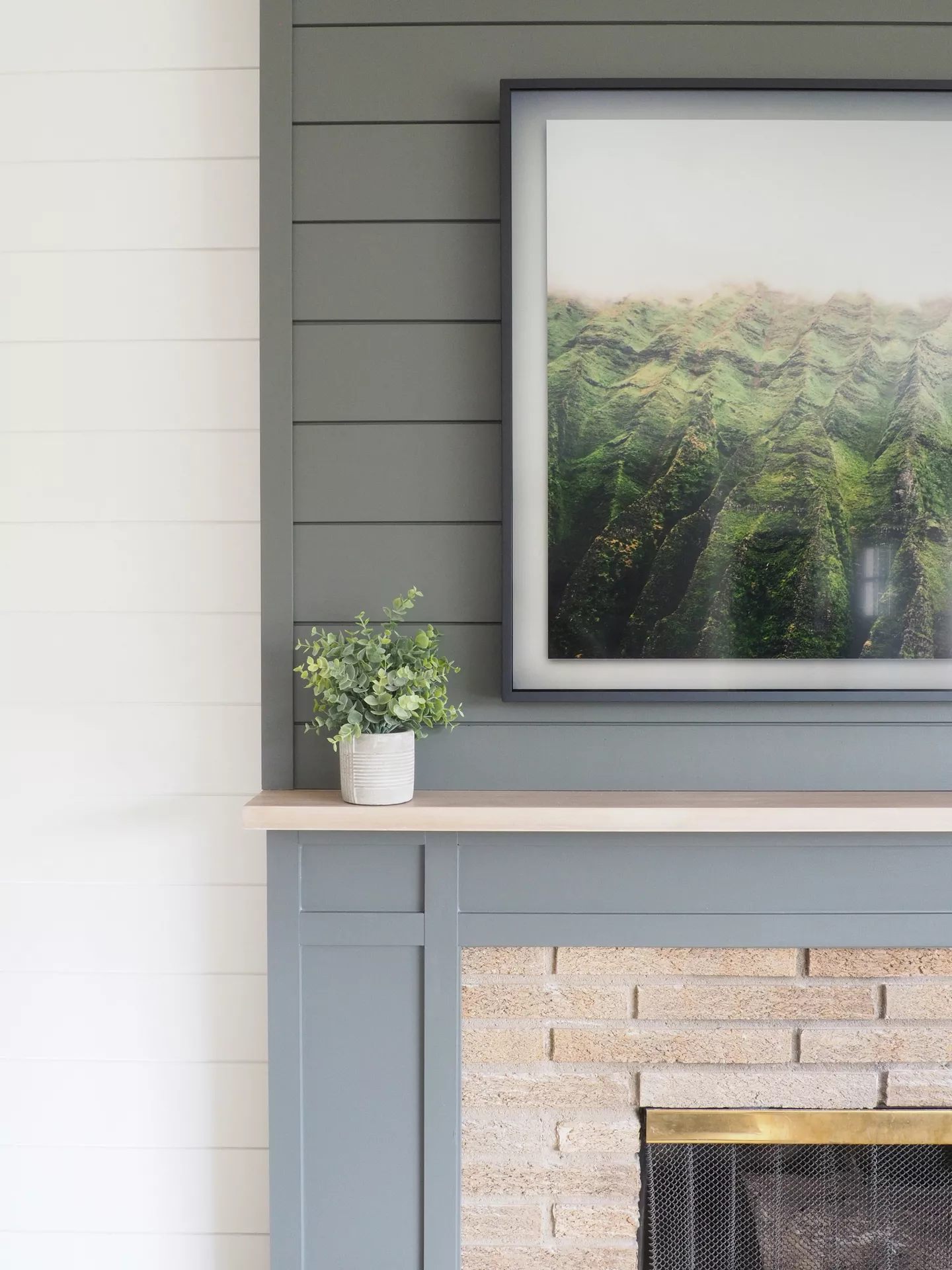 A modern fireplace with a slate-grey mantel and matching shiplap accent wall, featuring organic summer mantel ideas. Above the light-colored wooden shelf hangs a large, black-framed photograph depicting misty green mountain ridges, bringing nature based elements into the living space. A small, lush green plant in a white textured pot sits on the mantel, adding a touch of life against the muted grey and tan brickwork of the fireplace surround. The clean lines and serene imagery create a refreshing and contemporary seasonal display.