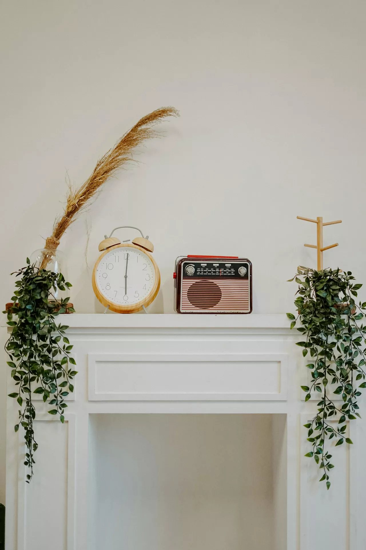 A clean white fireplace mantel showcases charming summer mantel ideas with a focus on vintage charm. The display features a collection of old school accents, including a large wooden alarm clock and a classic burgundy transistor radio positioned in the center. On either side, lush green artificial vines drape elegantly over the edges of the mantel. A tall sprig of dried pampas grass in a glass vase adds height on the left, while a minimalist wooden jewelry stand sits on the right, all set against a simple off-white wall for a nostalgic yet fresh seasonal look.