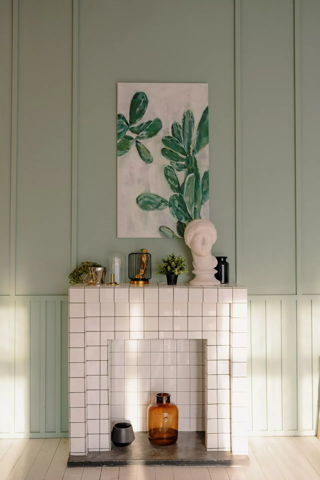 A modern fireplace tiled in white square ceramic serves as a bright base for eclectic summer mantel ideas. The arrangement includes a curated mix of plants, statue and gold accents, featuring a classical white bust, a small potted green plant, and a golden bird figurine inside a black wire cage. Above the mantel, a vertical canvas painting of a green cactus complements the pale sage-green paneled wall. Inside the fireplace hearth, a large amber glass bottle and a black ceramic bowl add additional texture to the sun-drenched, contemporary interior.