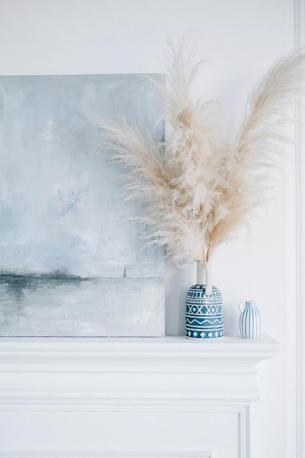 A serene close-up of a white mantelpiece showcasing coastal-inspired summer mantel ideas. An abstract canvas painting in shades of pale blue and grey leans against the wall, depicting a misty, ethereal landscape. Next to the artwork, a small blue and white ceramic vase with geometric patterns holds a large, fluffy arrangement of pampas grass, contributing to the room's soft vibes. A smaller, striped blue and white jug sits beside it, completing the bright and airy seasonal display.