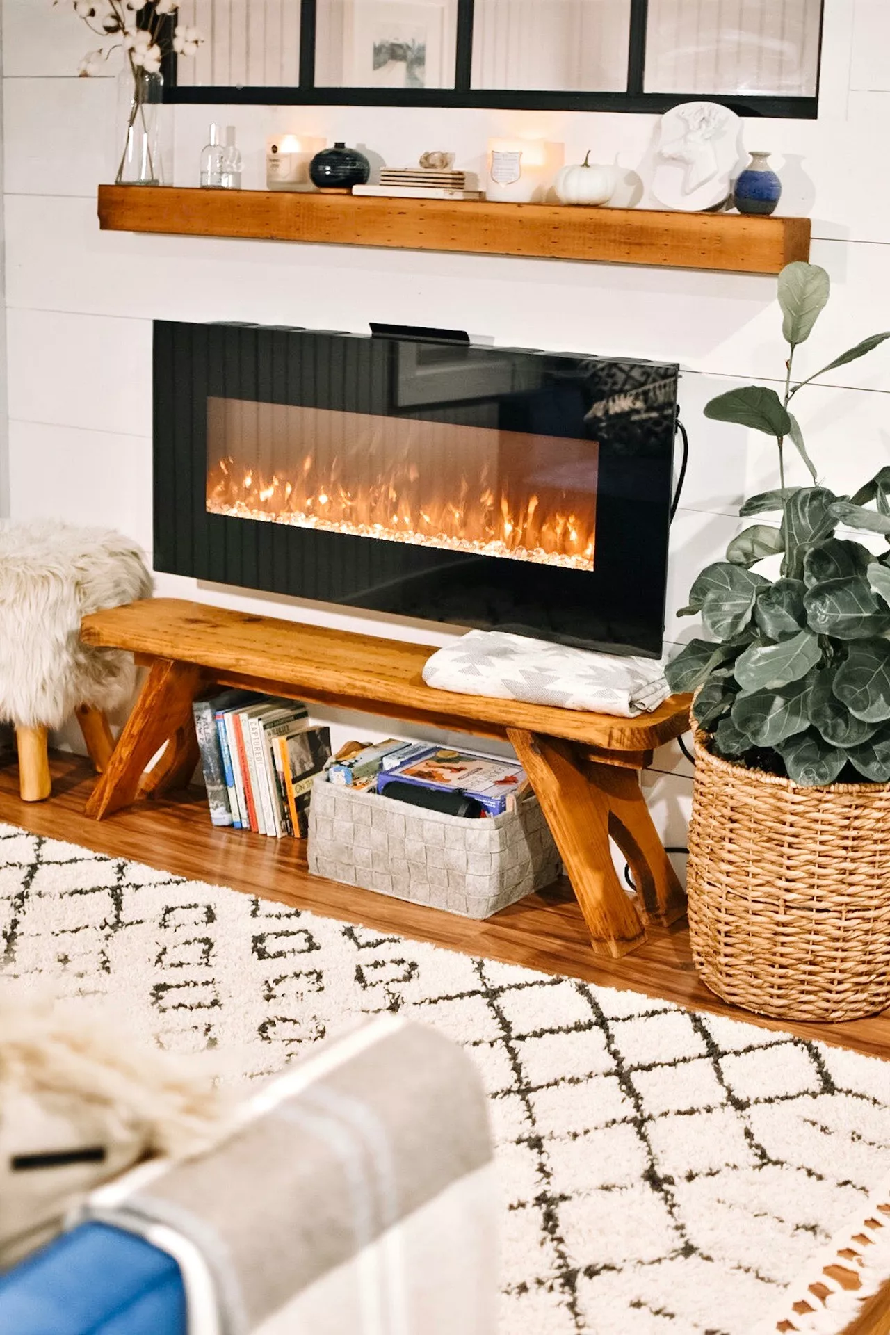 A modern living area featuring a sleek, wall-mounted electric fireplace set against white shiplap walls, showcasing cozy summer mantel ideas. The floating wooden mantel above is decorated with various stylish accents, including glowing scented candles, small ceramic vases in navy and white, and a decorative white pumpkin. Below the fireplace, a rustic wooden bench holds a folded grey patterned throw, while a grey felt basket and a collection of books are tucked neatly underneath. A large woven basket with a lush green plant and a patterned black-and-white diamond rug complete the comfortable, contemporary interior.
