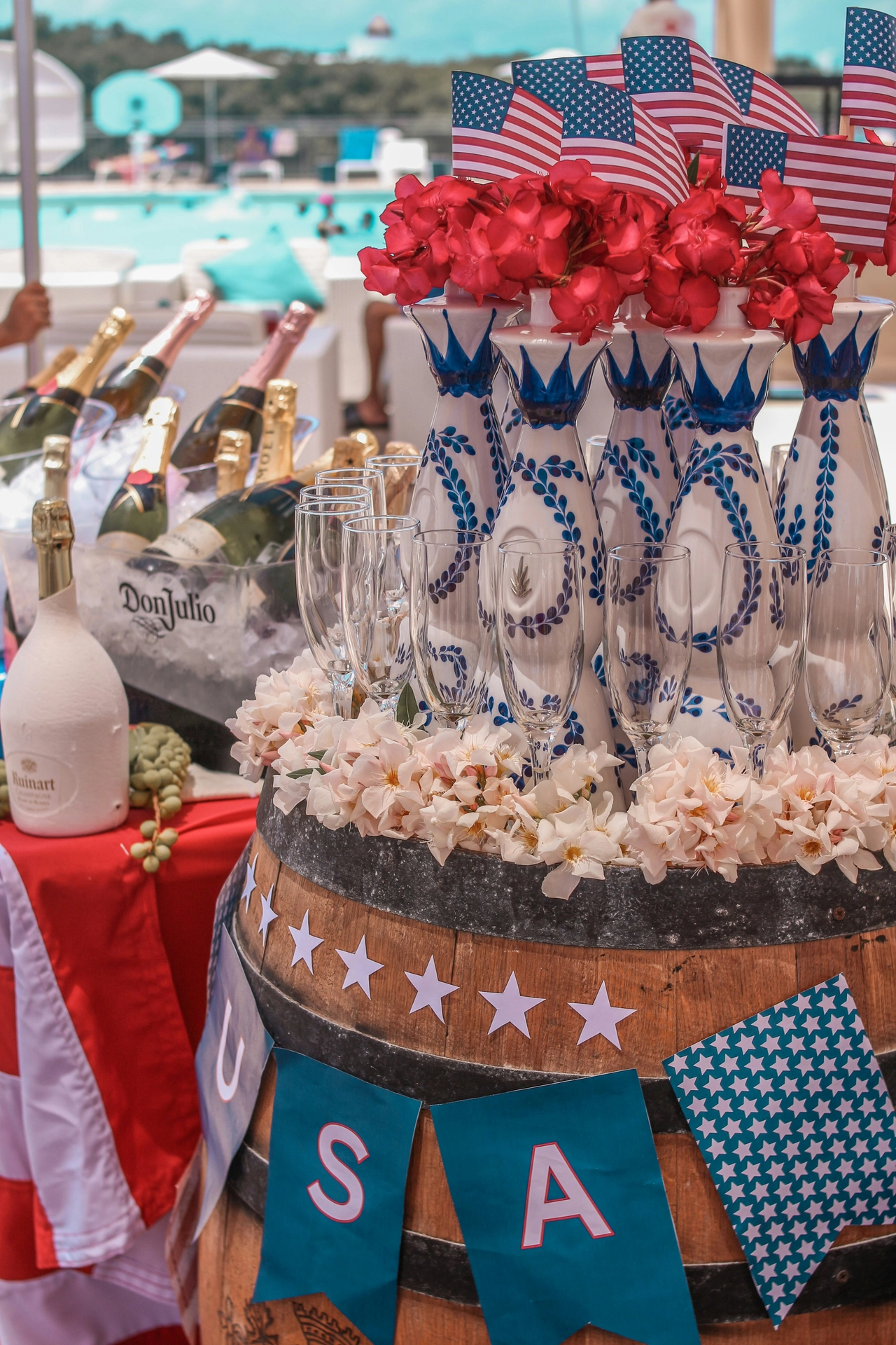A festive poolside beverage station highlights patriotic 4 july decor arranged on a rustic wooden barrel. Serving as vibrant 4th of july party decor, the top of the barrel is lined with clear champagne flutes, white floral garlands, and distinctive blue-and-white patterned ceramic bottles topped with bright red flowers and small American flags. A teal pennant banner spelling out 