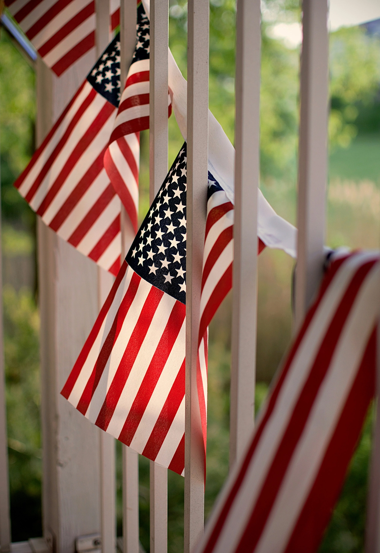 A close-up, angled view captures classic 4 july decor woven through an outdoor porch railing. A festive flag garland made of consecutive small American flags is draped horizontally between the white vertical slats of the deck balustrade. The bright summer sunlight filters through the fabric of the flags, highlighting the red and white stripes and the blue field of white stars. The soft-focus green background of trees and a lawn enhances the warm, patriotic, and celebratory backyard atmosphere.