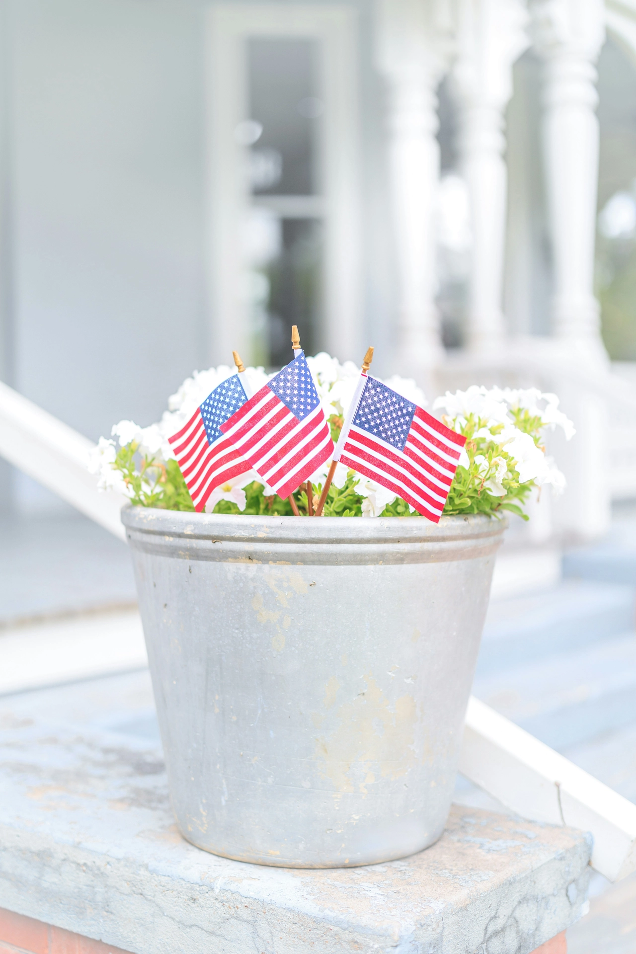 A charming outdoor setting features simple festive 4 july decor on a porch entrance. A vintage, weathered silver metal bucket is used as a planter, displaying small American flags in flower pot arrangements tucked neatly into a cluster of blooming white flowers. The bucket rests on a light blue painted concrete step next to a white handrail. In the soft-focus background, the classic architecture of a white house porch with traditional columns enhances the bright, patriotic, and summery look of the display.