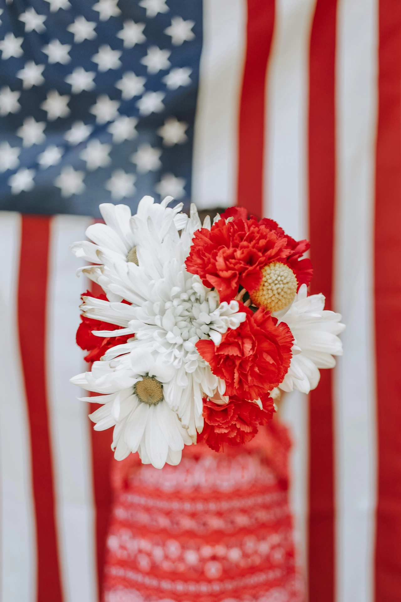 A vibrant and patriotic close-up captures festive 4 july decor with a beautiful hand-held bouquet. The arrangement features fresh flowers, including large white daisies, a textured white chrysanthemum, and ruffled bright red carnations, creating a classic festive color scheme. In the background, a large American flag with its iconic stars and stripes hangs vertically, providing a bold patriotic backdrop. A person wearing a red patterned top or dress is softly blurred behind the floral arrangement, making the bouquet the striking central focal point of the celebratory scene.