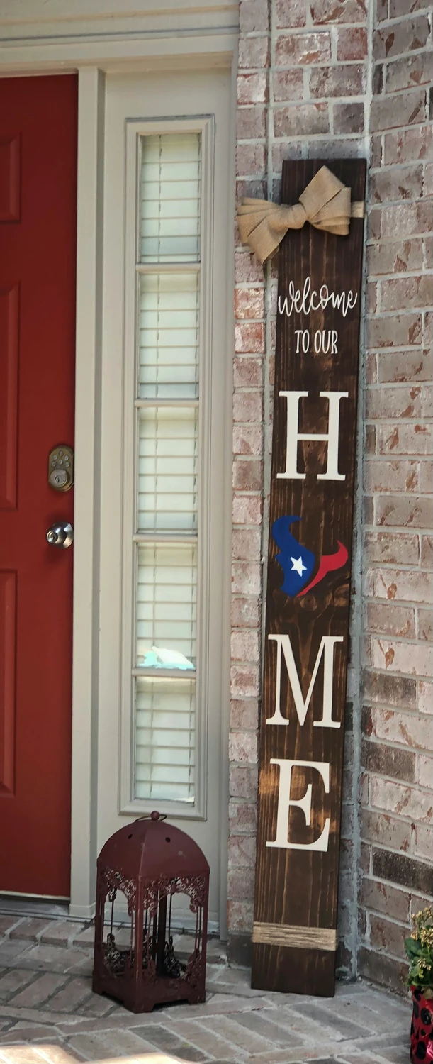 A welcoming entryway features patriotic 4 july decor displayed alongside a red door to create a classic front door decor style. Leaning against a red brick wall is a tall, dark stained vertical wooden sign reading 