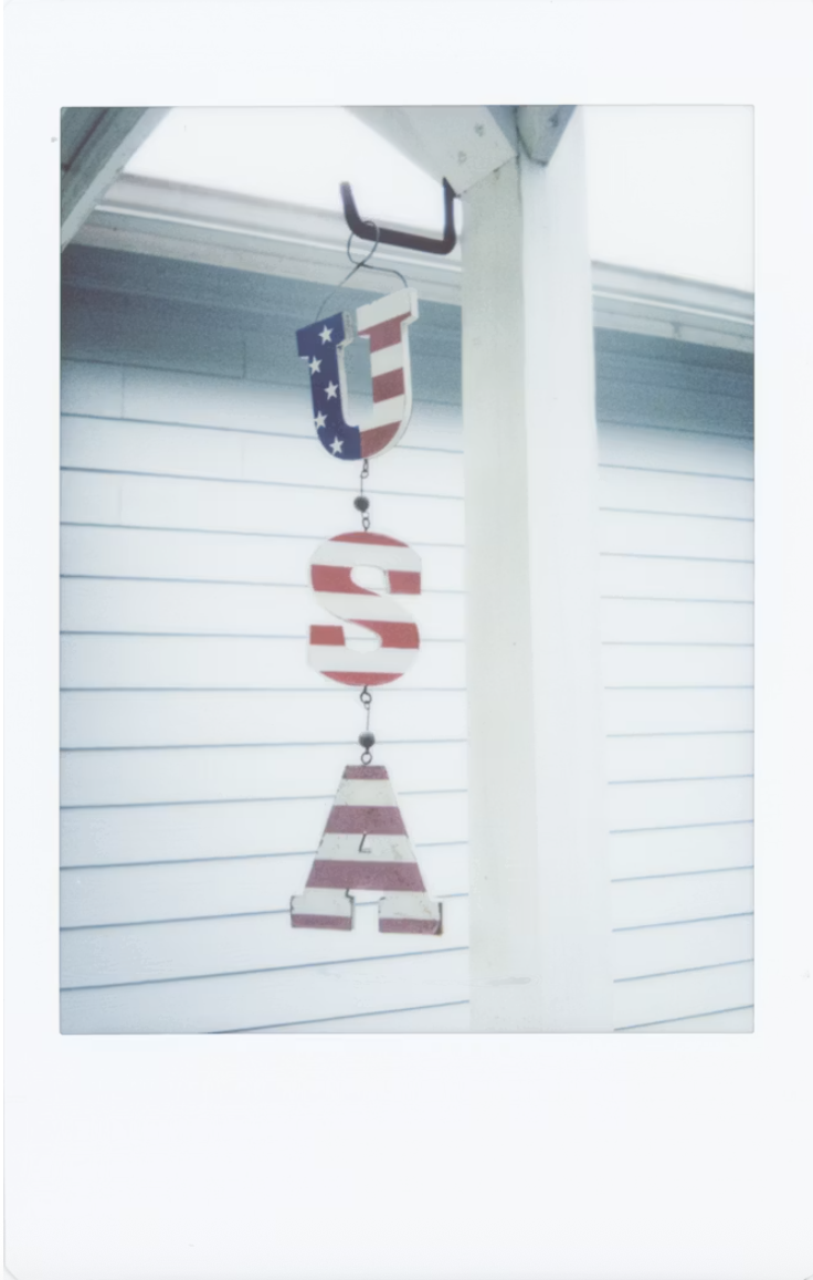 A nostalgic, Polaroid-style photograph captures rustic 4 july decor hanging outdoors. Suspended vertically from a black metal hook on a white porch beam is a decorative chain of wooden letters spelling out 