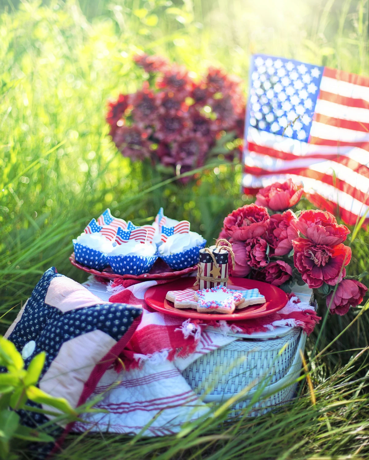 A festive outdoor setup hidden in tall green grass showcases creative 4 july decor arranged on a white woven picnic basket. Serving as a charming picnic decor idea, the top of the basket is layered with a red-and-white checked cloth, a star-shaped patriotic patchwork pillow, and red plates displaying frosted cupcakes with small American flags and star-shaped sugar cookies. A bouquet of ruffled deep red flowers sits beside the treats, while a full-sized American flag and additional red blossoms flutter softly in the sunlit, blurry background, creating a whimsical and patriotic summer field celebration.