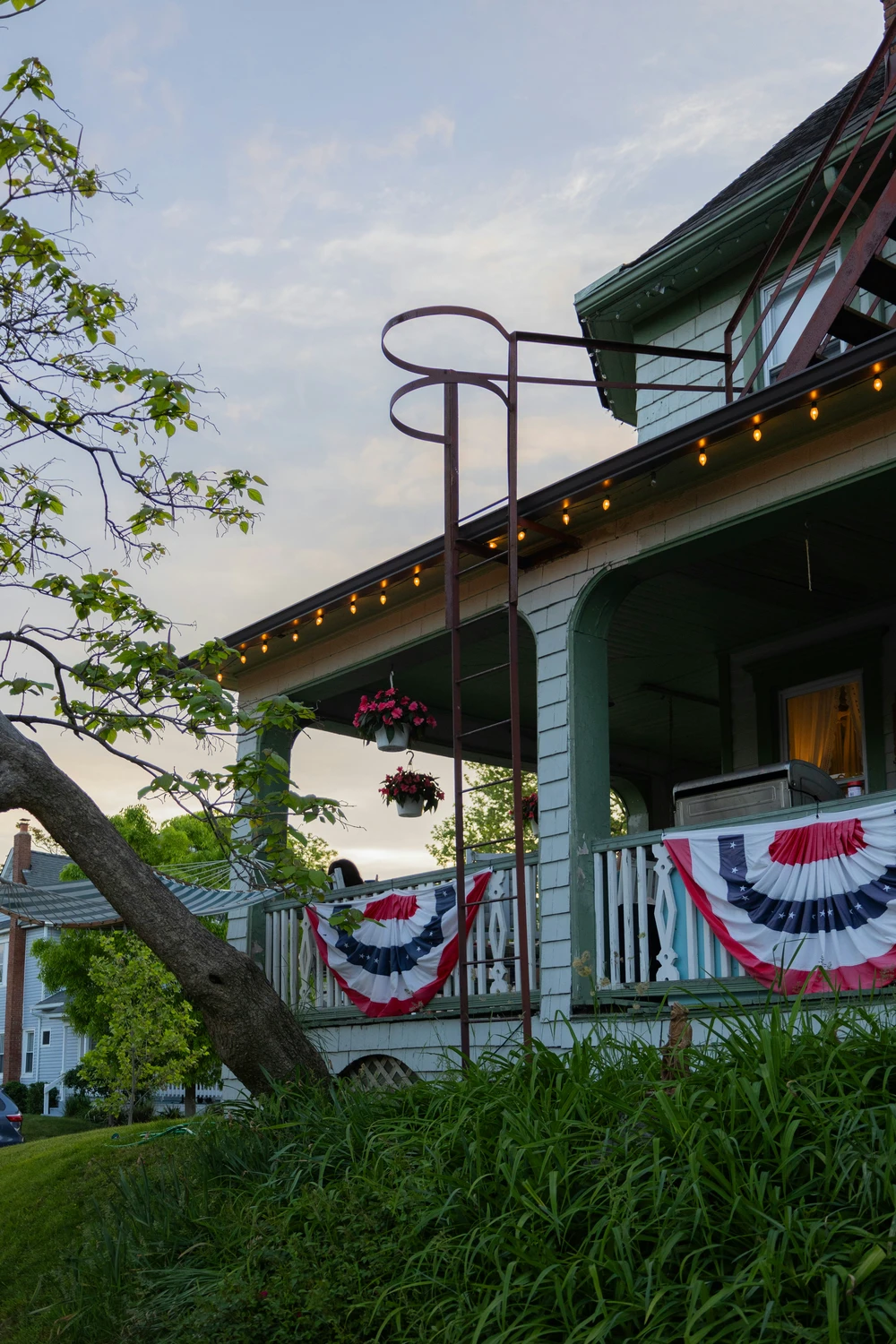 A charming, green-trimmed historic house displays festive 4 july decor along its spacious wrap-around porch. Hanging prominently from the white wooden porch railing is a classic pleated fan flag bunting with red, white, and blue stripes adorned with stars. Warm string lights glow softly under the porch eaves, complemented by hanging flower baskets with red blooms. In the foreground, a grassy hill with lush green foliage and a leaning tree frames the house, while a metal fire escape ladder extends upward against a pale evening sky.